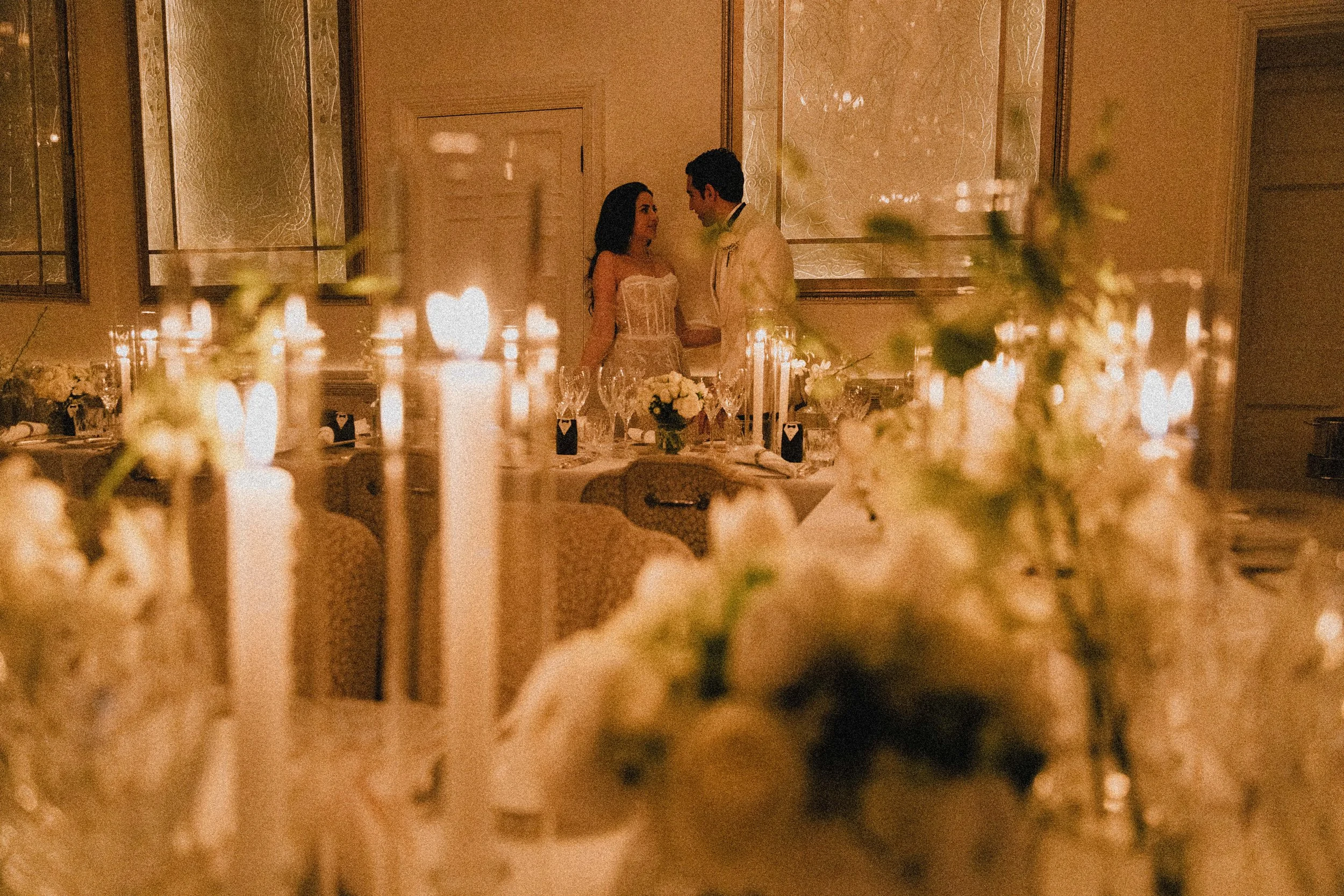 A couple in wedding attire dancing or sharing an intimate moment in a warmly lit reception hall decorated with candles and floral arrangements.