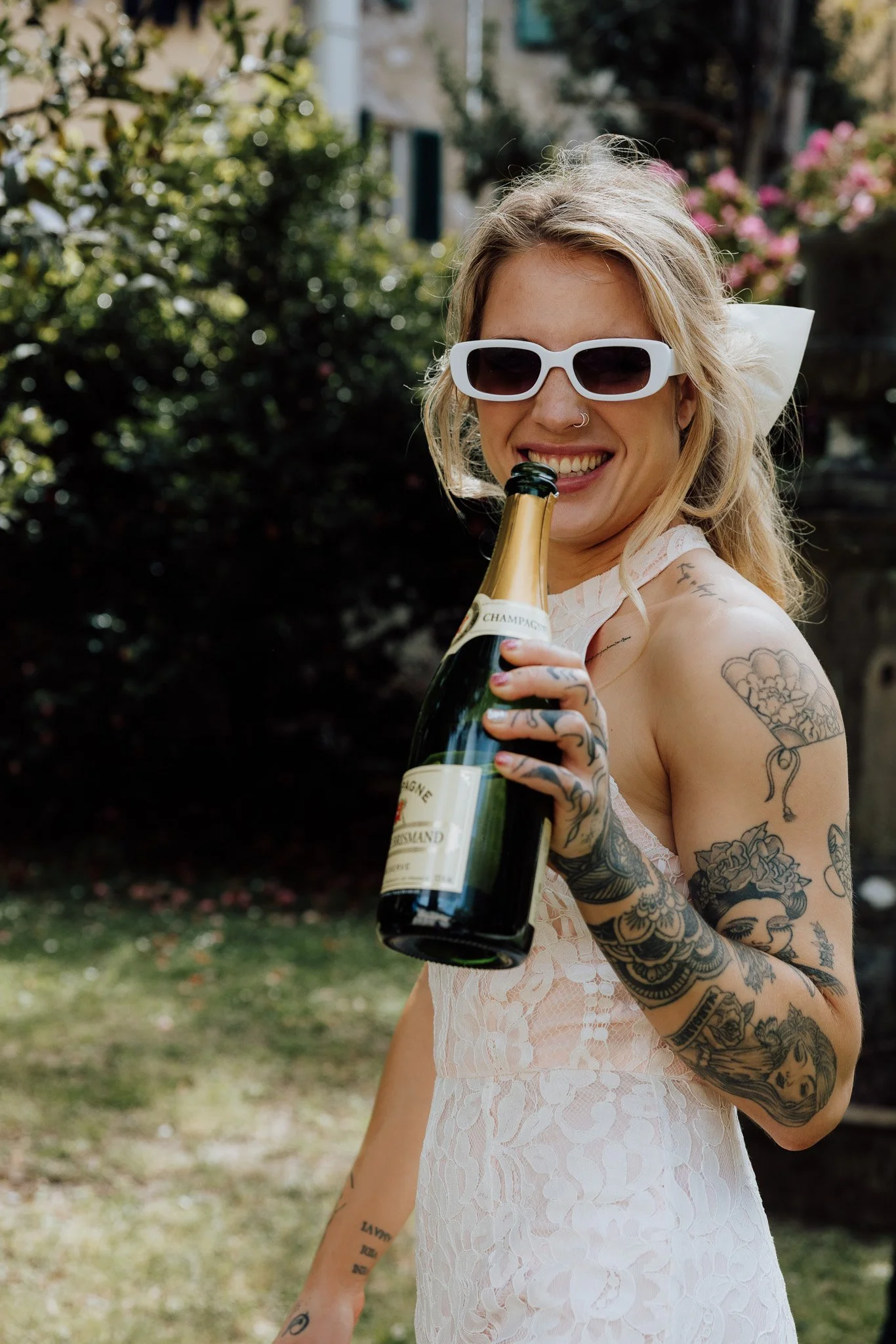 A woman with tattoos and blonde hair, wearing white sunglasses and a lace dress, holding a bottle of champagne outdoors.