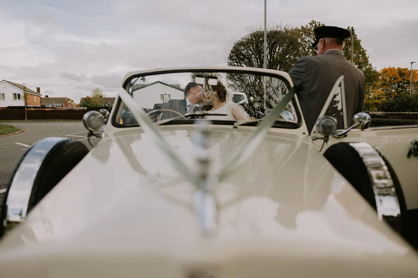 A wedding couple sitting inside a vintage convertible car, seen through the front of the vehicle, with a man standing outside.