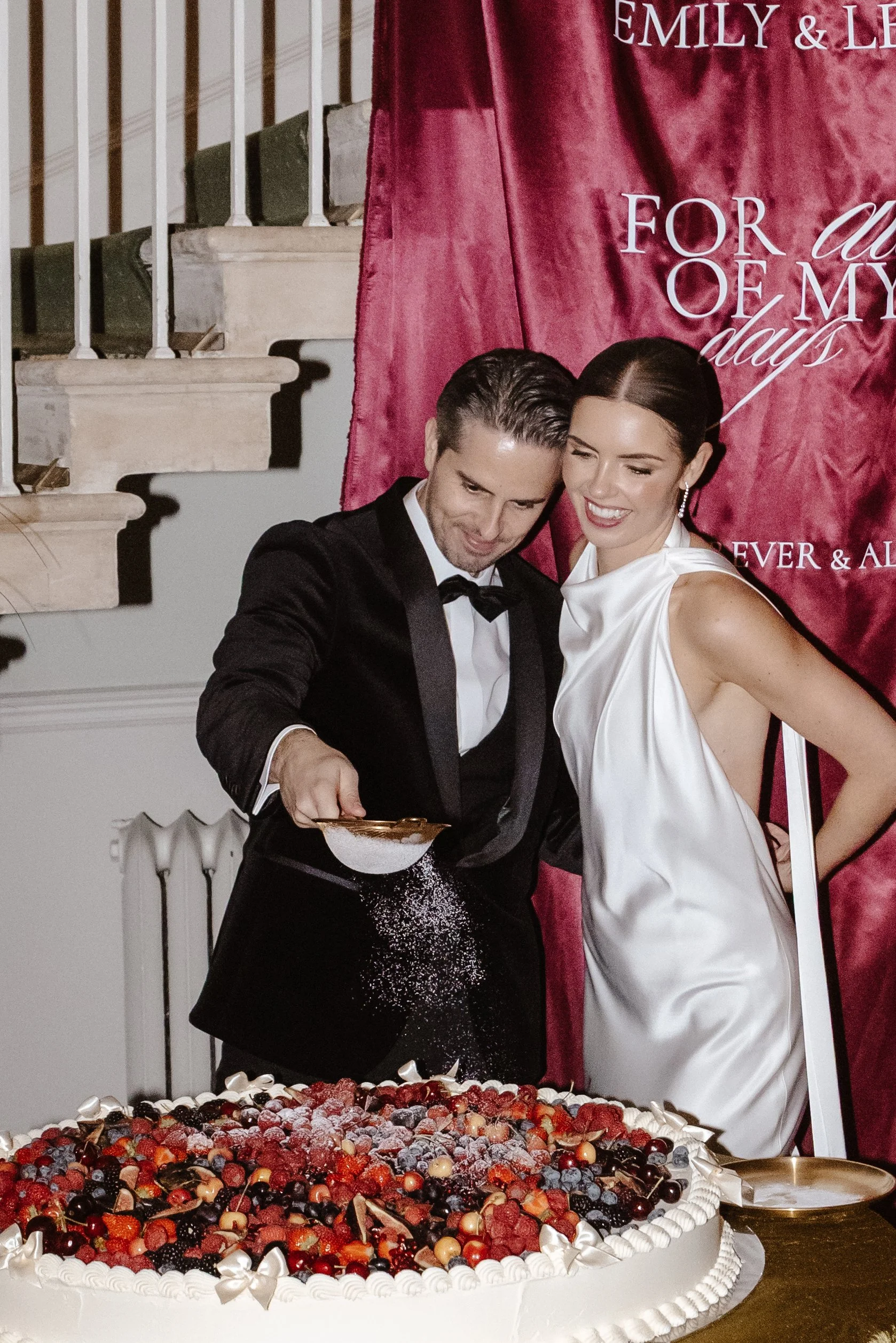 A couple dressed in formal attire, the man in a tuxedo and the woman in a white satin gown, smiling as the man sprinkles powdered sugar onto a large decorated cake during a celebration. middleton lodge weddings 