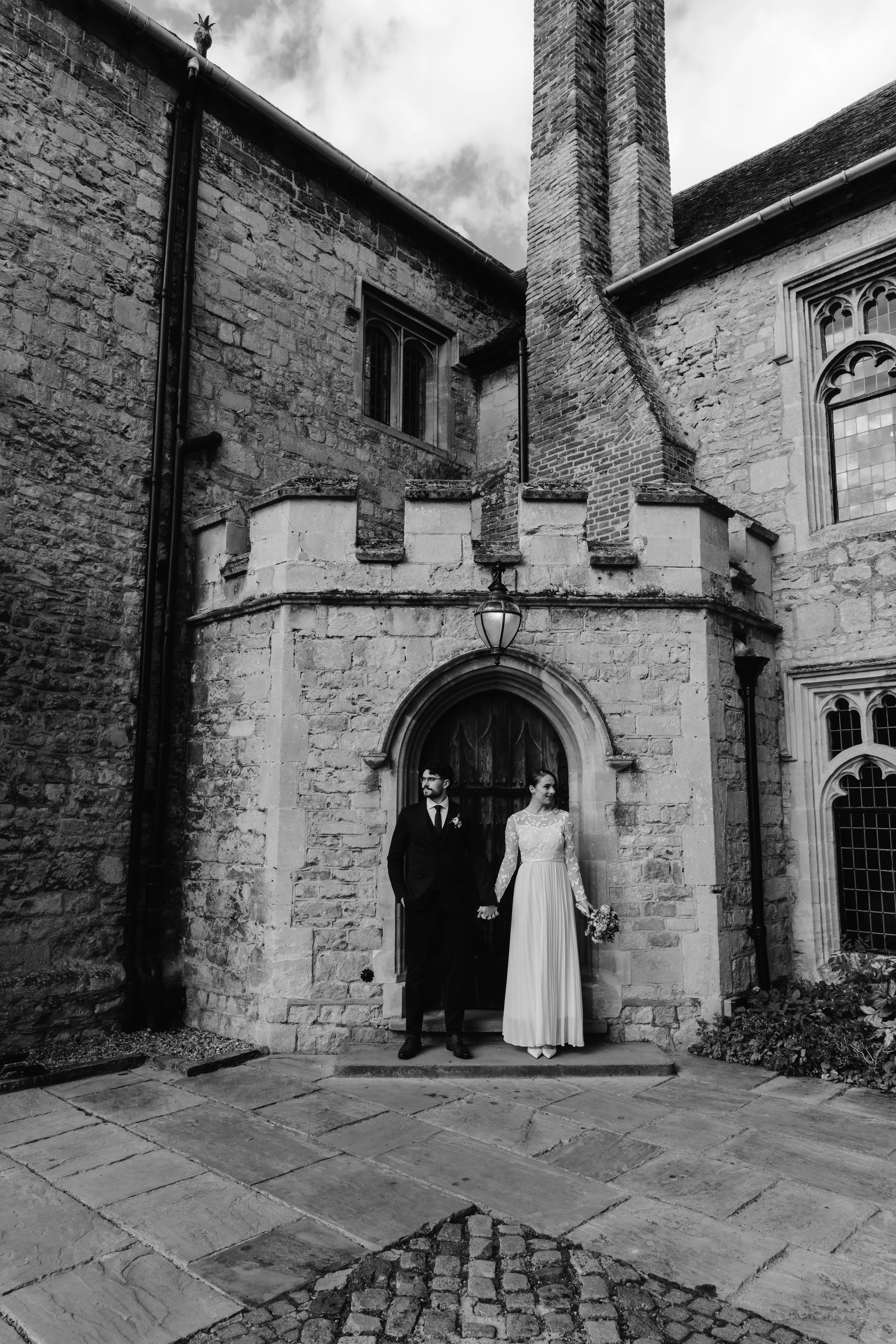 A couple in wedding attire standing outside a historic stone church, holding hands and looking in different directions, with a cloudy sky overhead. wedding in york castle 
