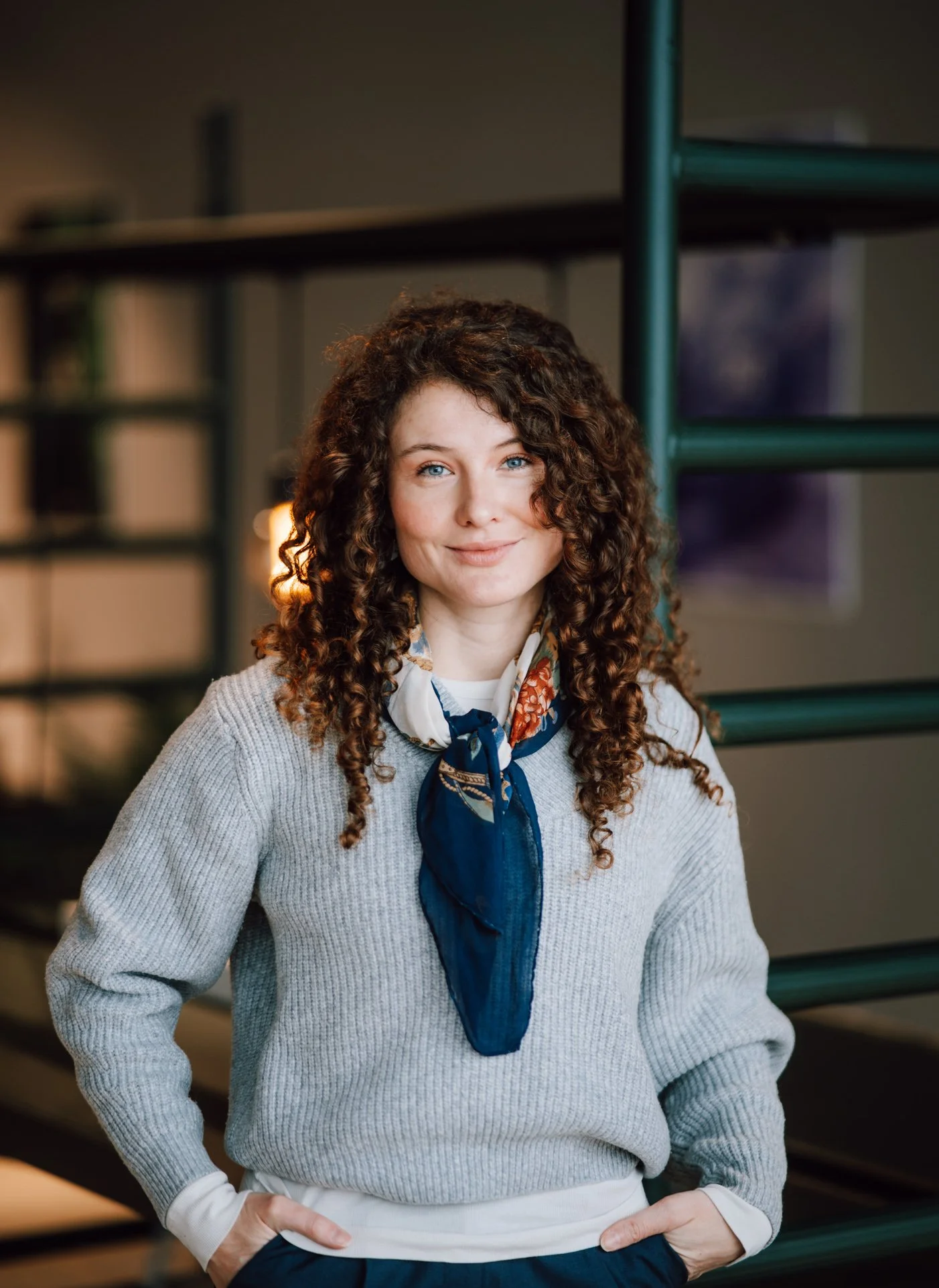 A woman with curly brown hair and blue eyes wearing a gray sweater and a colorful scarf, standing indoors with her hands in her pockets and smiling gently.