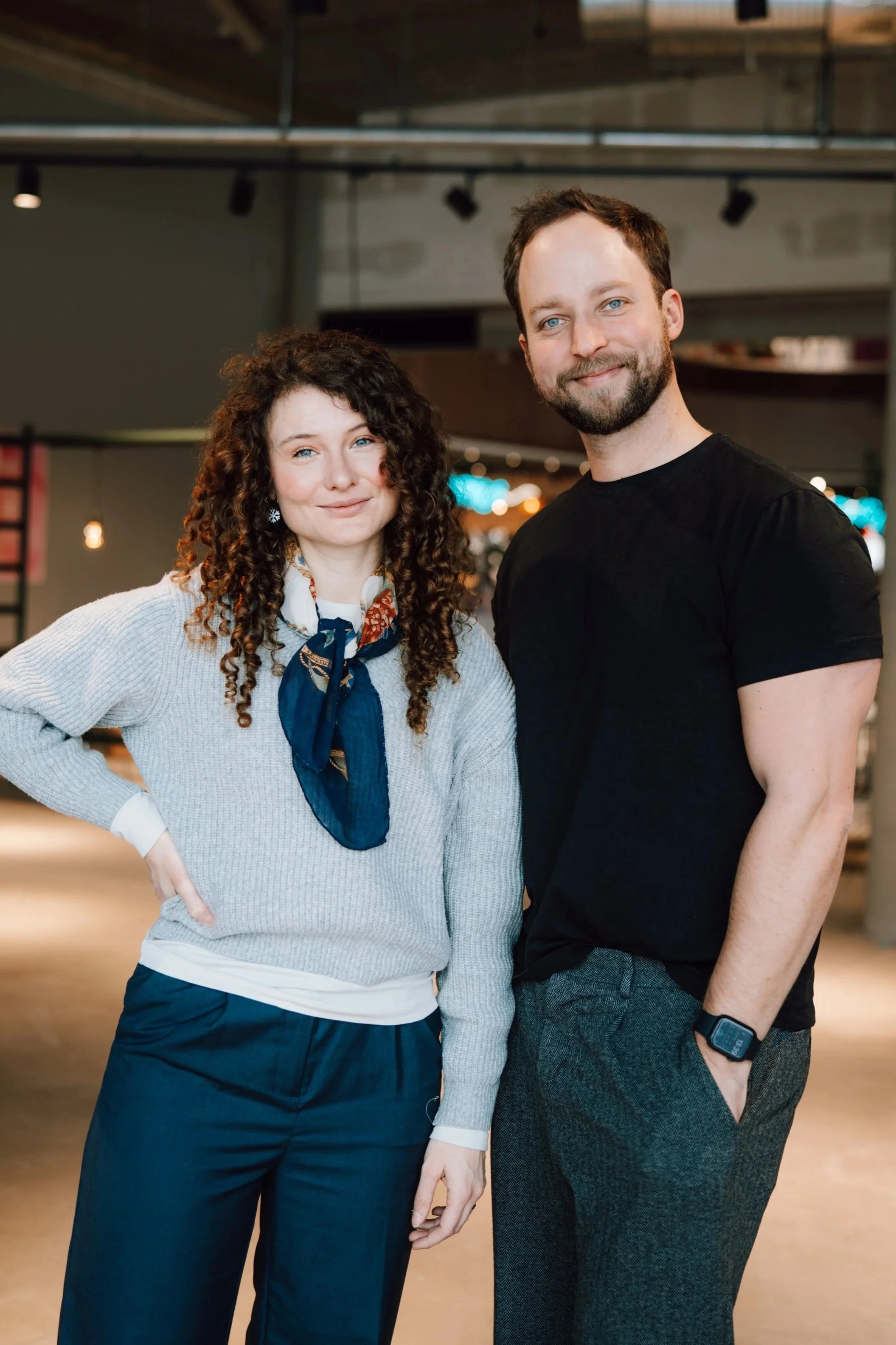 A woman and a man standing indoors, smiling at the camera. The woman has curly hair, wearing a gray sweater with a blue scarf, and dark pants. The man has a beard, wearing a black t-shirt, gray pants, and a smartwatch on his wrist.