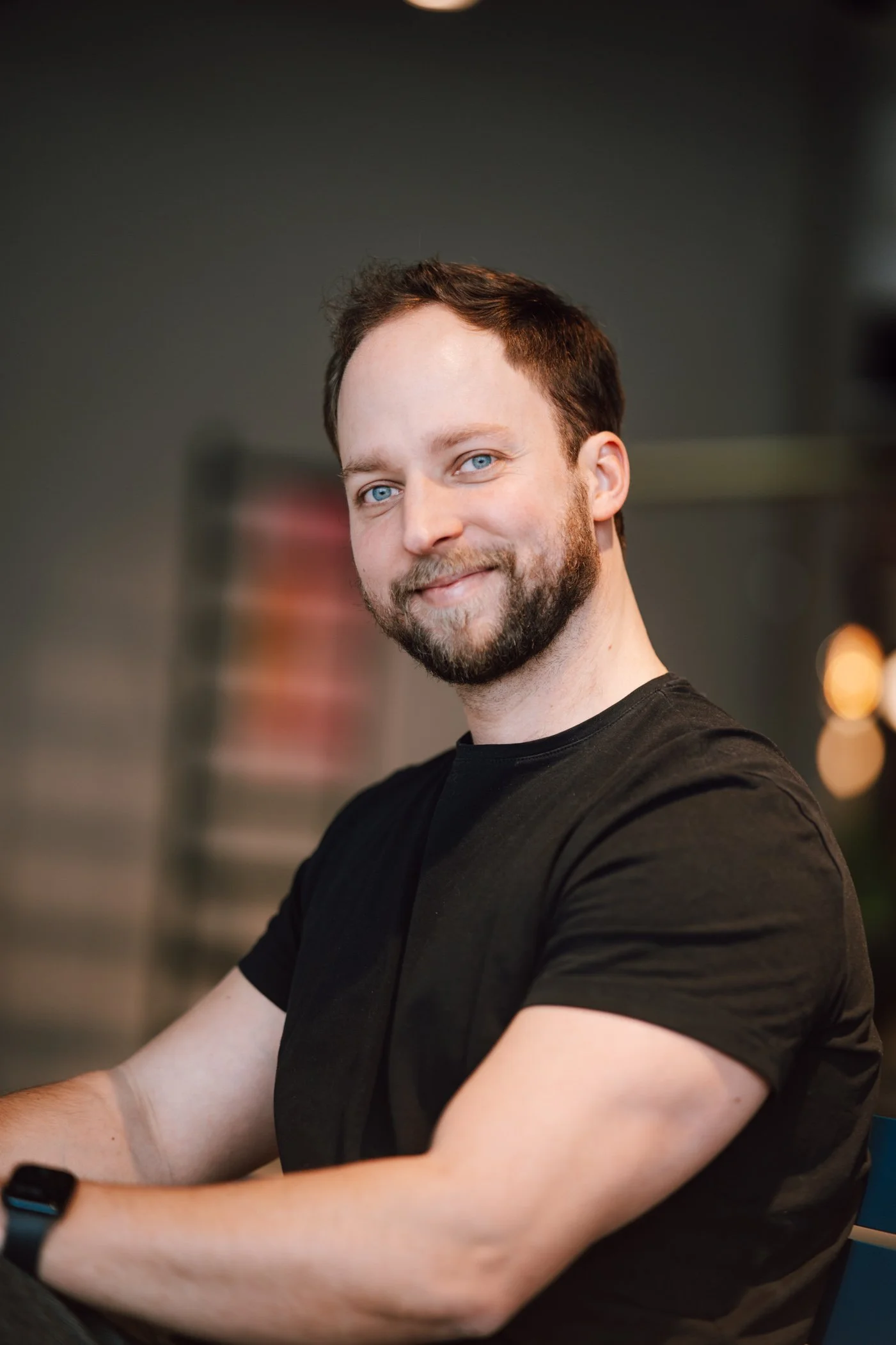 Portrait of a smiling man with blue eyes and brown hair, wearing a black t-shirt, sitting indoors.