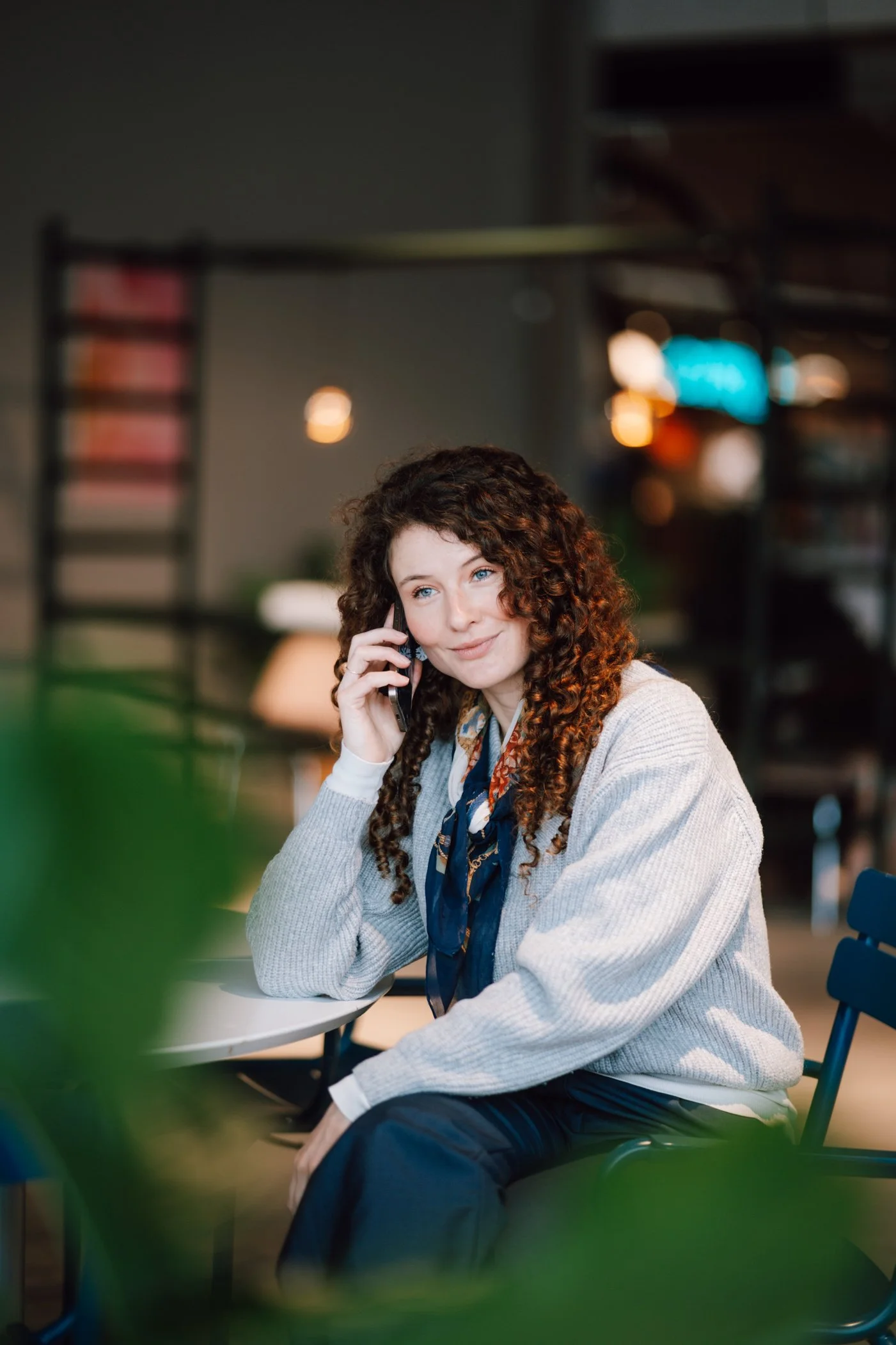 A woman with curly brown hair and blue eyes sitting at a table, talking on her cell phone in a cafe or restaurant with warm lighting and blurred background.