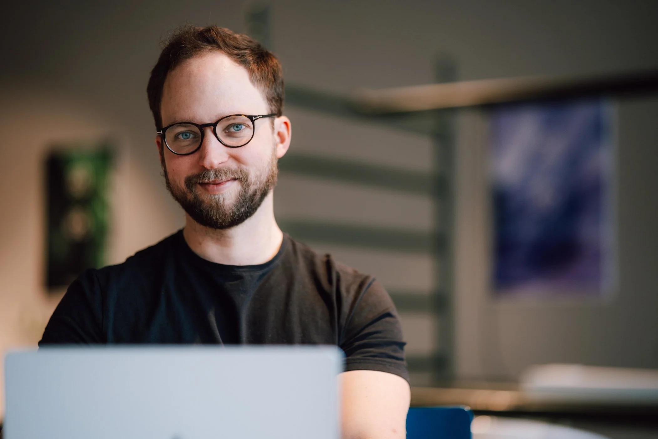 Smiling man with glasses and a beard working on a laptop in an office or workspace.