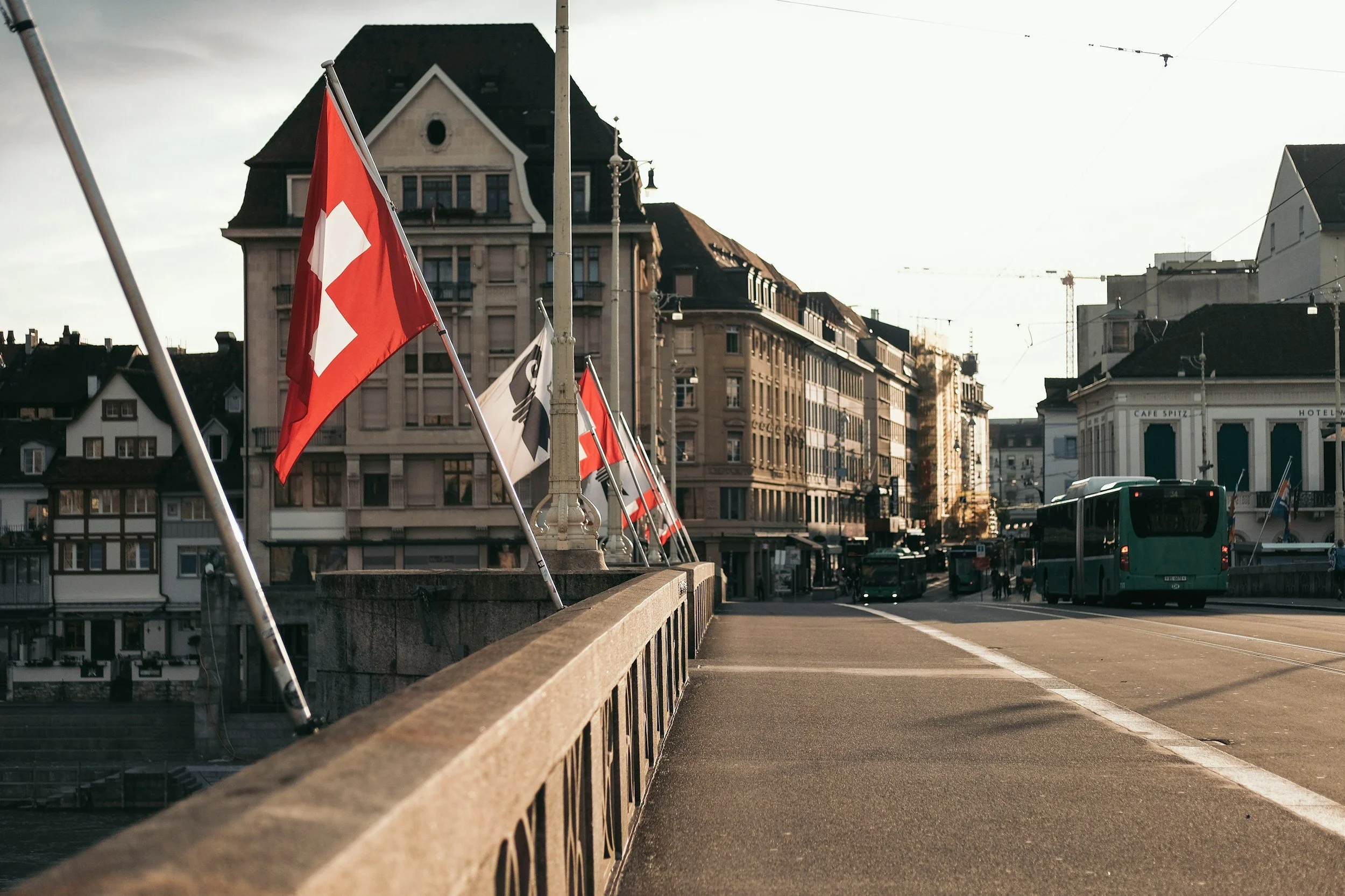 Stadtansicht mit Schweizer Flaggen auf einer Brücke, im Hintergrund alte Gebäude und Busse auf der Straße.