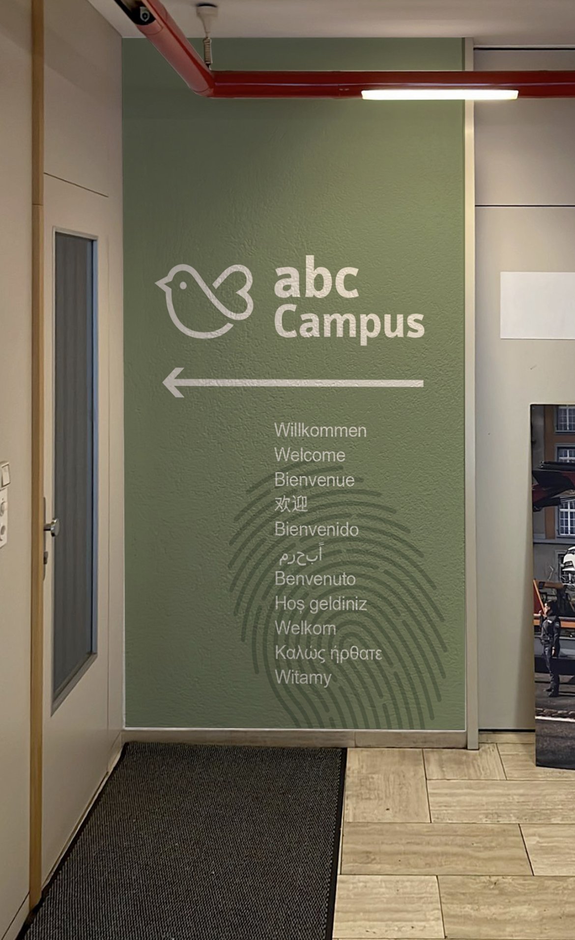 Wand mit Willkommensgruß in mehreren Sprachen und Spielfeder-Logo für den ABC Campus, Richtung links