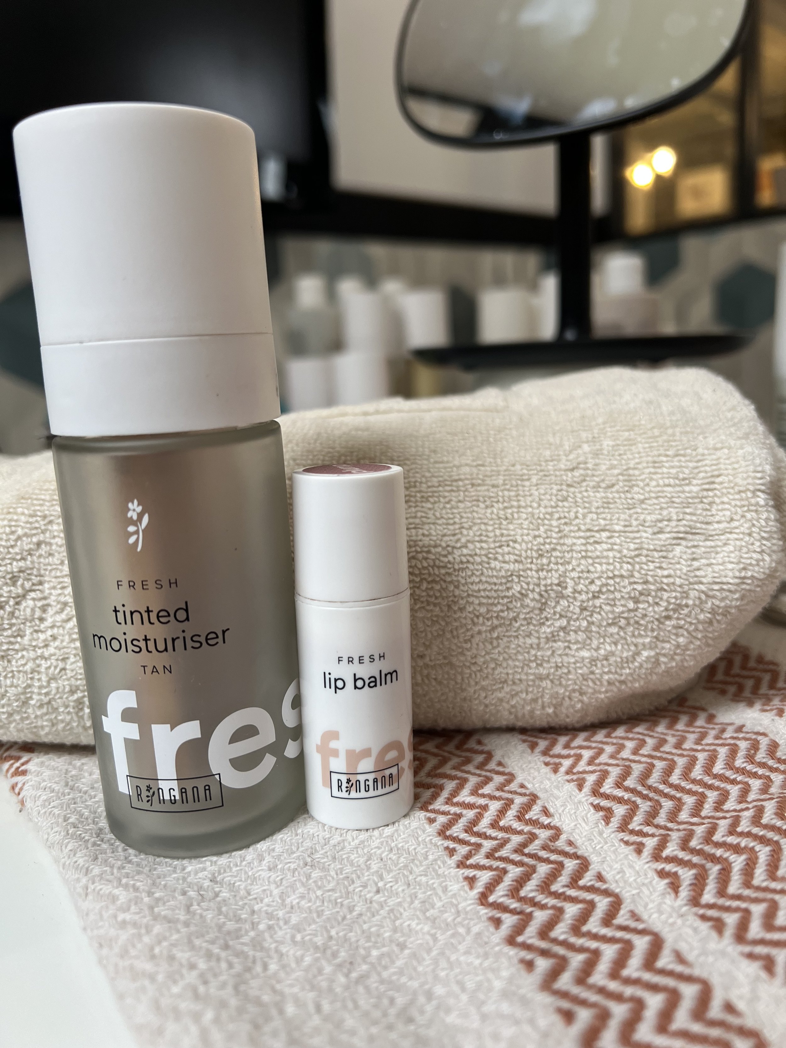 Deux produits de soins pour la peau, une crème teintée SPF et une baume à lèvres (lip balm), placés sur une serviette blanche, avec un miroir en arrière-plan.