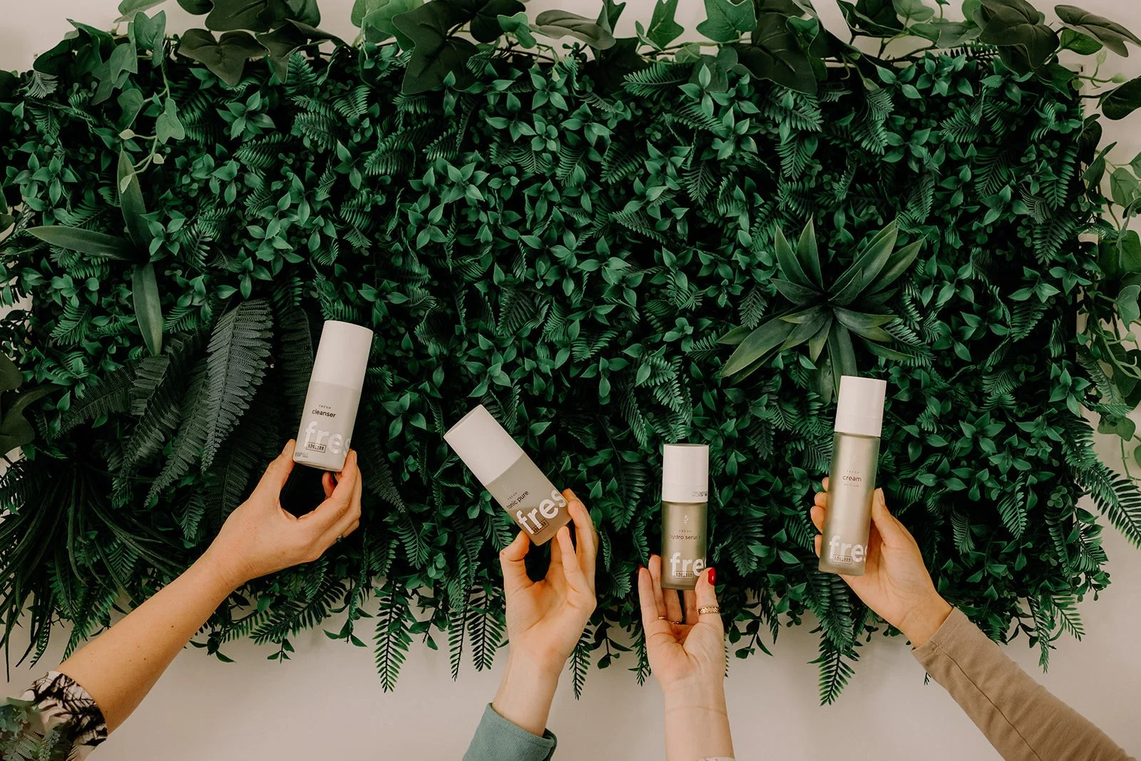 Quatre mains tenant des bouteilles de produits de soin ringana (soin naturel et frais) devant un mur de plantes vertes.