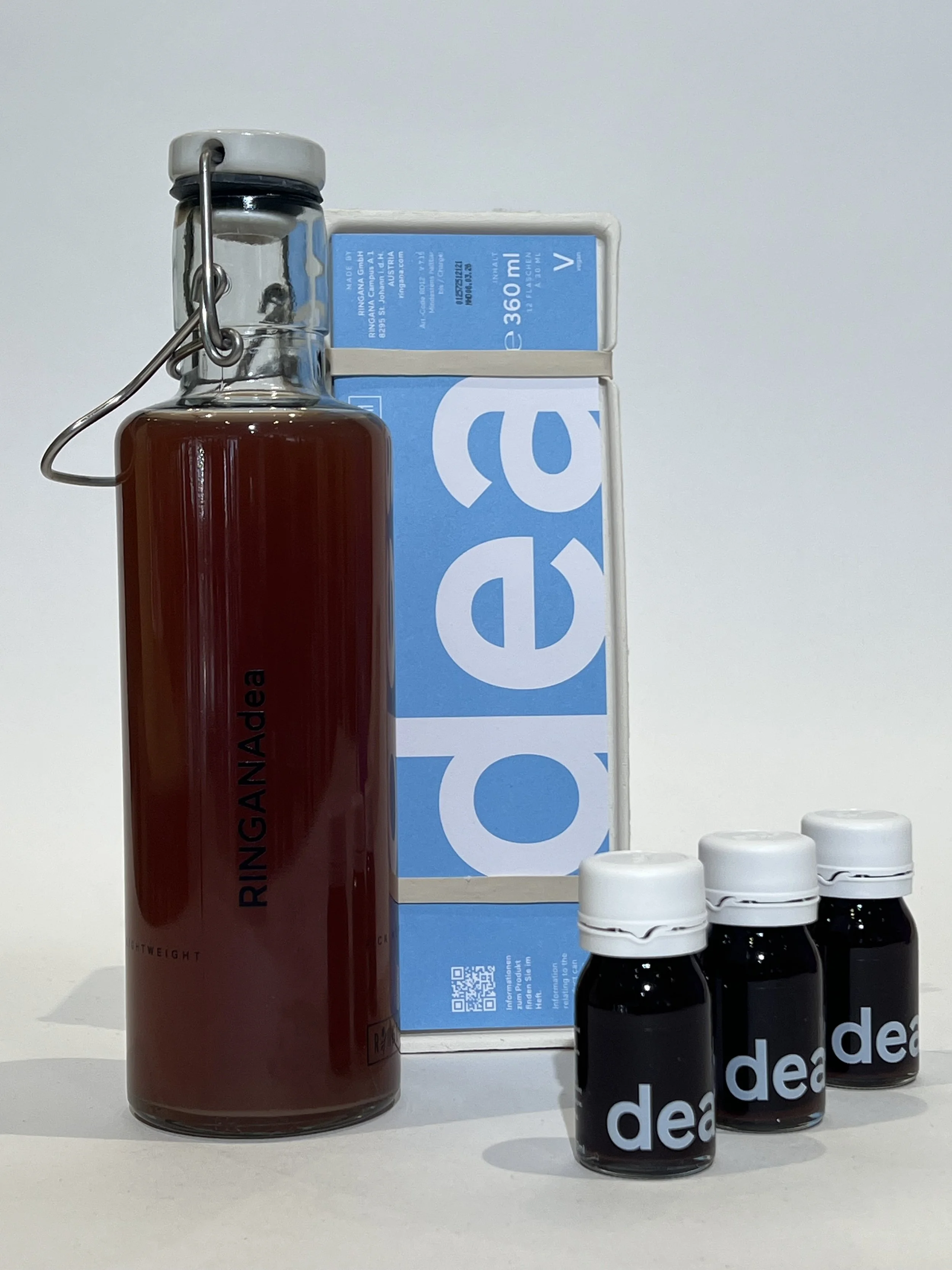 Une bouteille en verre avec un liquide rouge foncé, une boîte bleue et blanche, et trois petits flacons noirs avec bouchons blancs et le mot 'DEA' écrit dessus. boisson poids plume, drainante, perte de poids