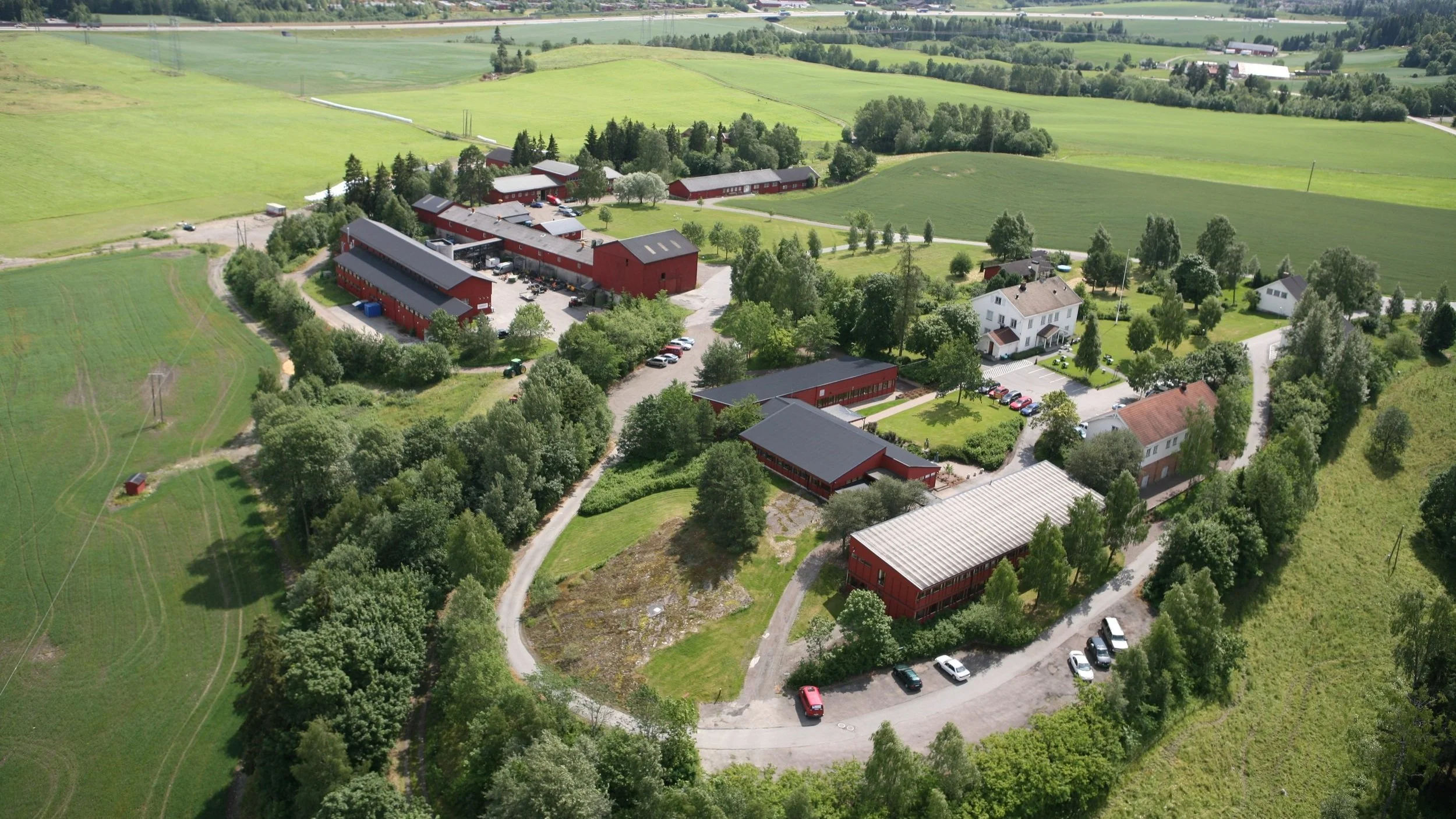 Landskap med grønne åkre, trær, røde bondegårder og bygninger, samt kjøretøy parkert langs veier.