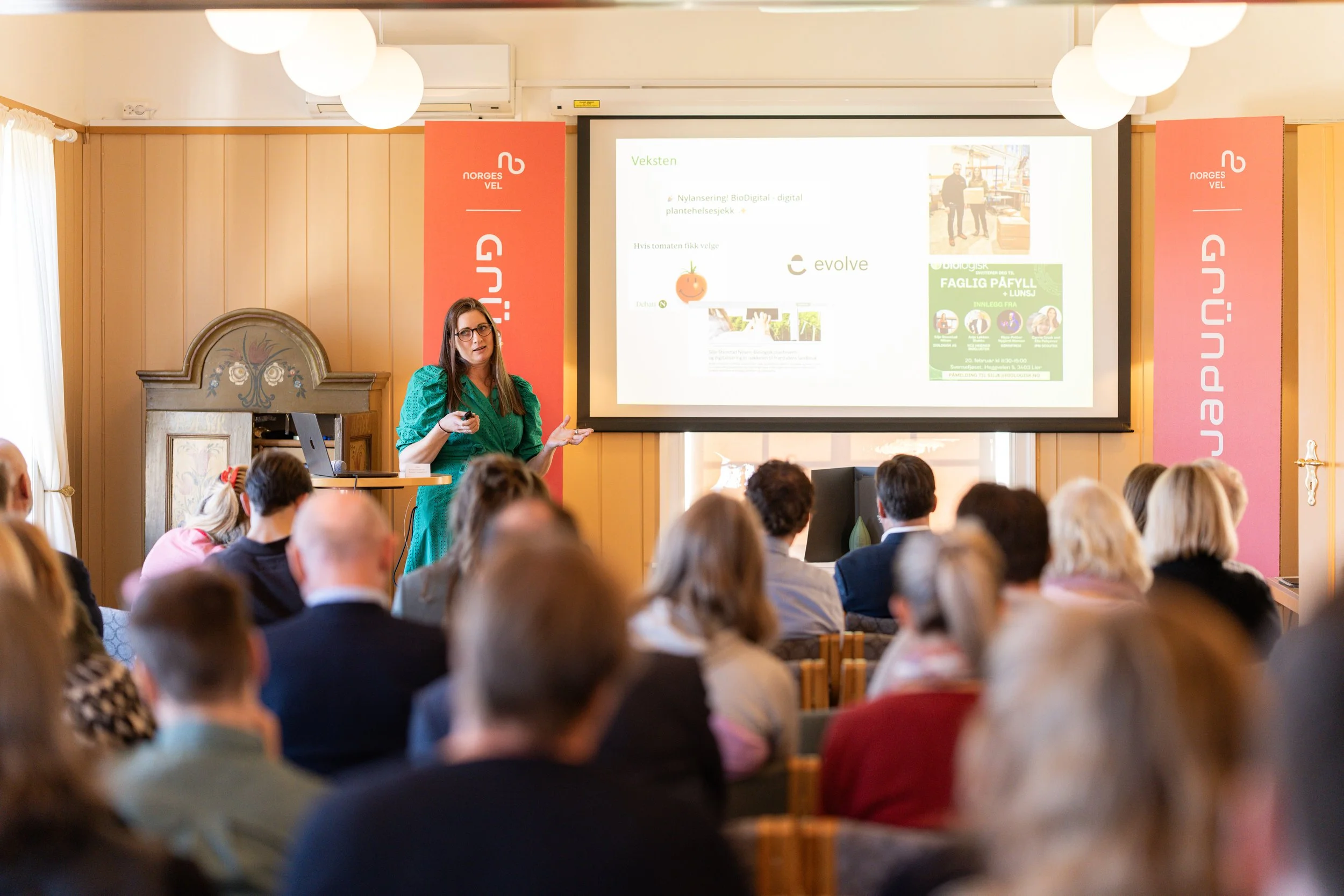 En kvinne holder en presentasjon foran et foran et publikum i et møterom, med en stor skjerm som viser lysbilde. Publikum består av voksne personer som sitter og lytter.