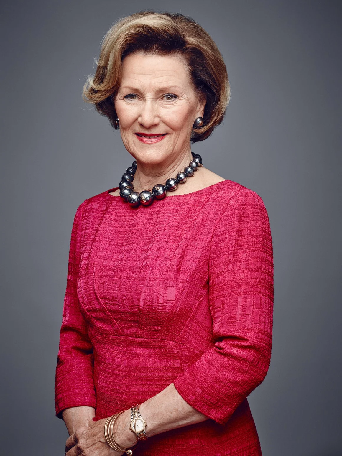 Portrett av Dronning Sonja, iført en rød kjole og perle- og gullakser, mot en grå bakgrunn.