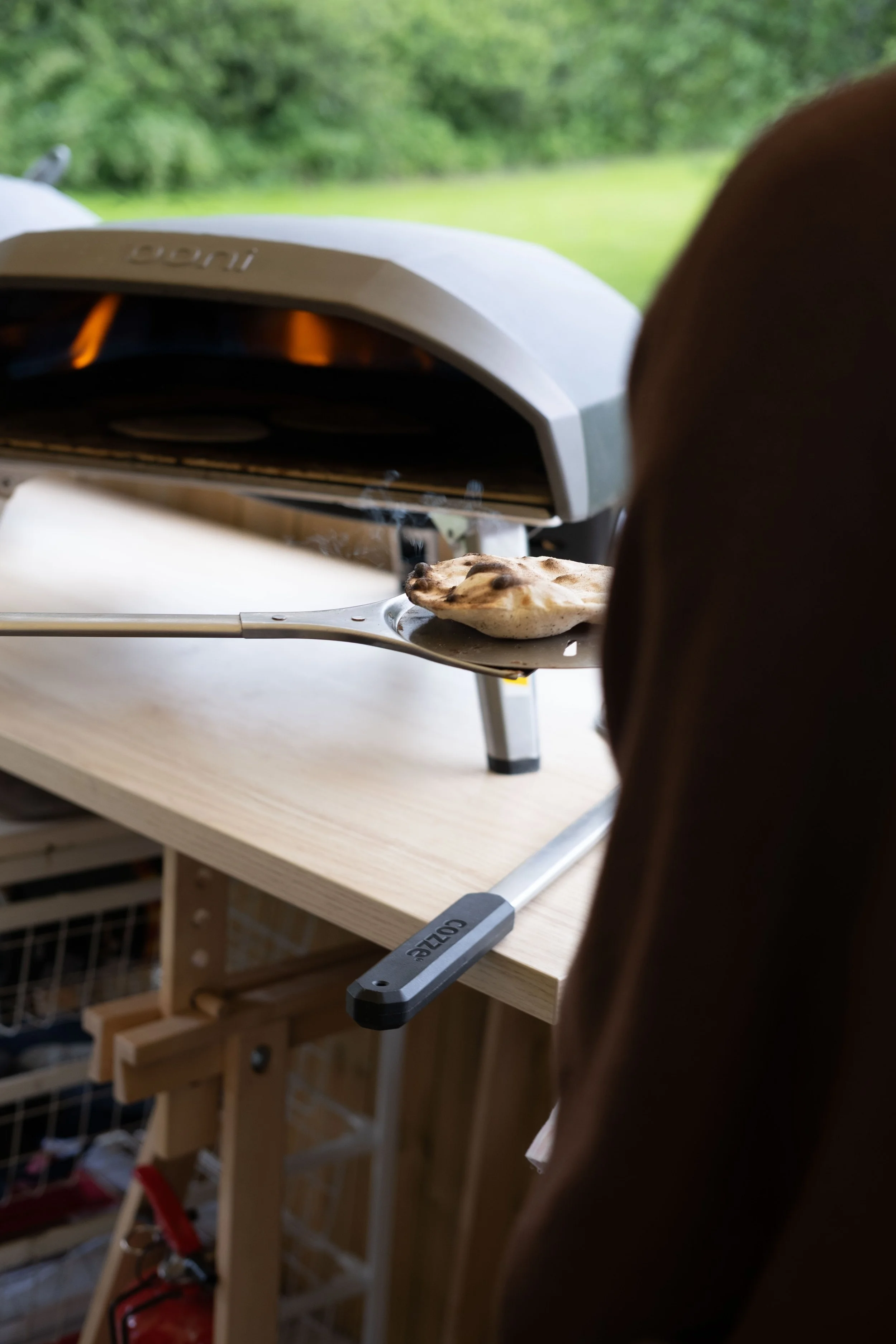 Et lite elektrisk grill med grillmat på en trebordplate i et rom med litt uklar utendørs bakgrunn.