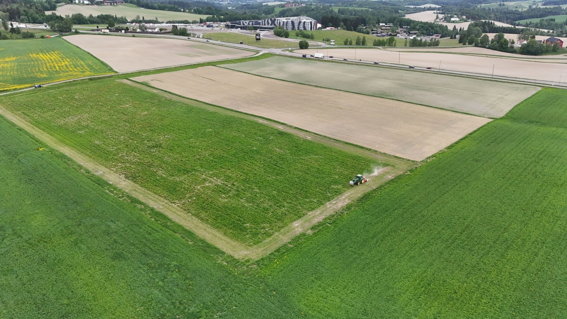 Landskap med grønne jordbruksfelt, en traktor som bearbeider jorden, og veier som går gjennom landskapet. Det er bygninger og skog i bakgrunnen.