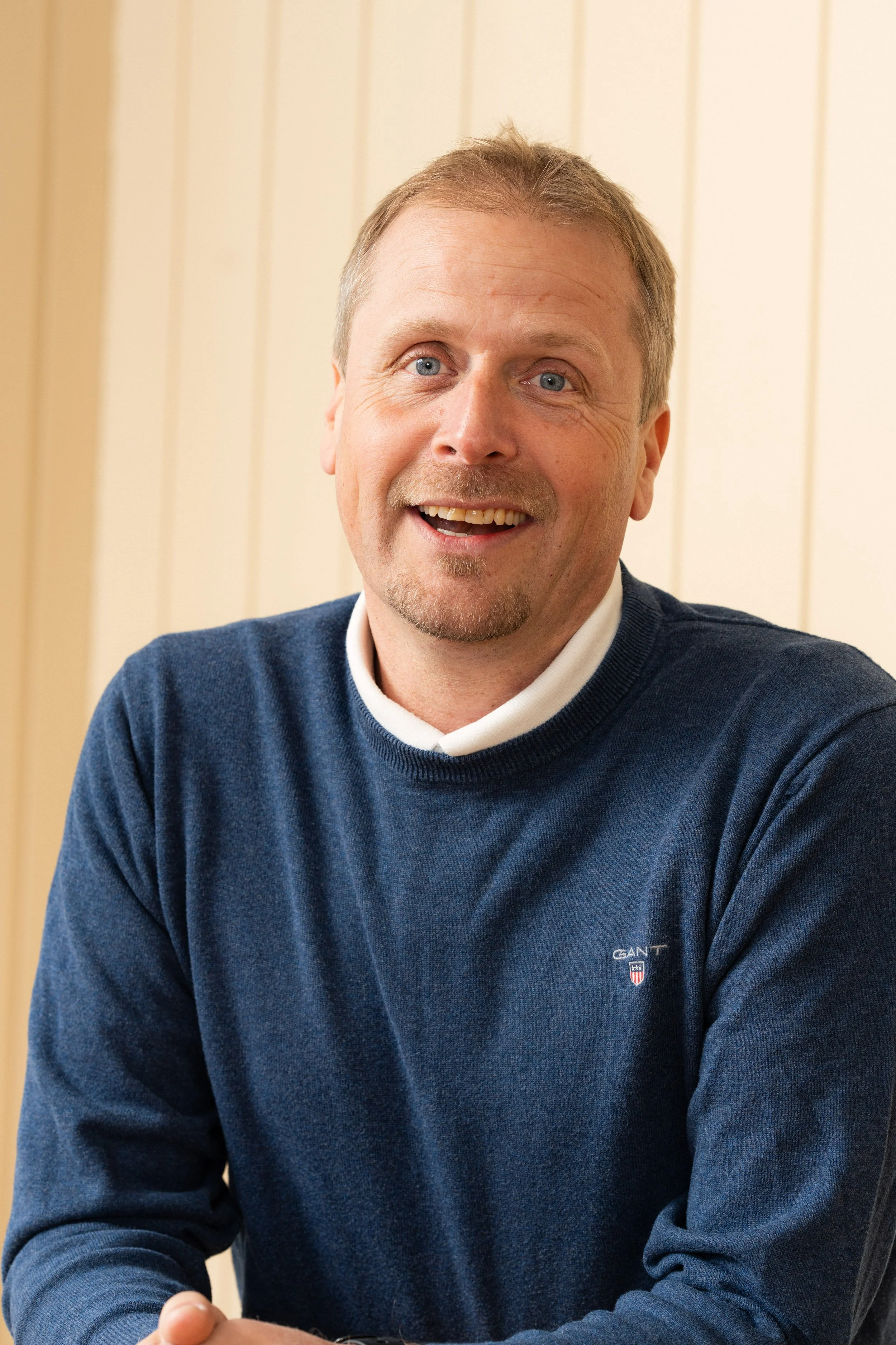 Et bilde av en smilende mann med blonde hår, blå øyne og skjegg, iført en blå genser med en liten logo på venstre bryst. Bakgrunnen består av en lysfarget, vertikal trepanel.