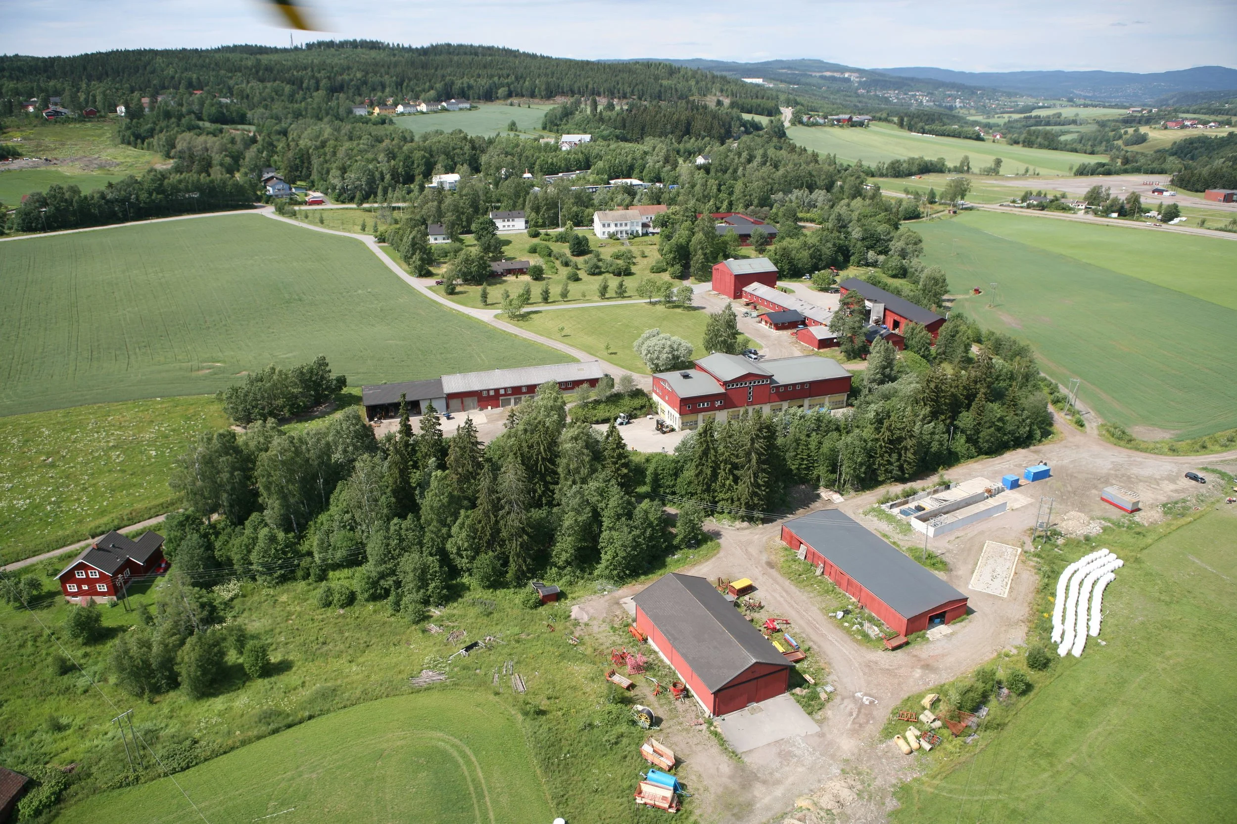 Landskap med gårdsbruk, grønne åkrer, skog og noen bygninger i en rural setting.