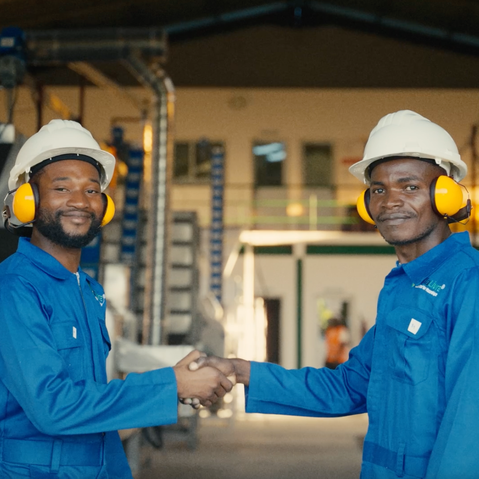 To menn i blå arbeidstøy, begge med hjelm og hørselsvern, håndhilser i en fabrikk eller industrihall.