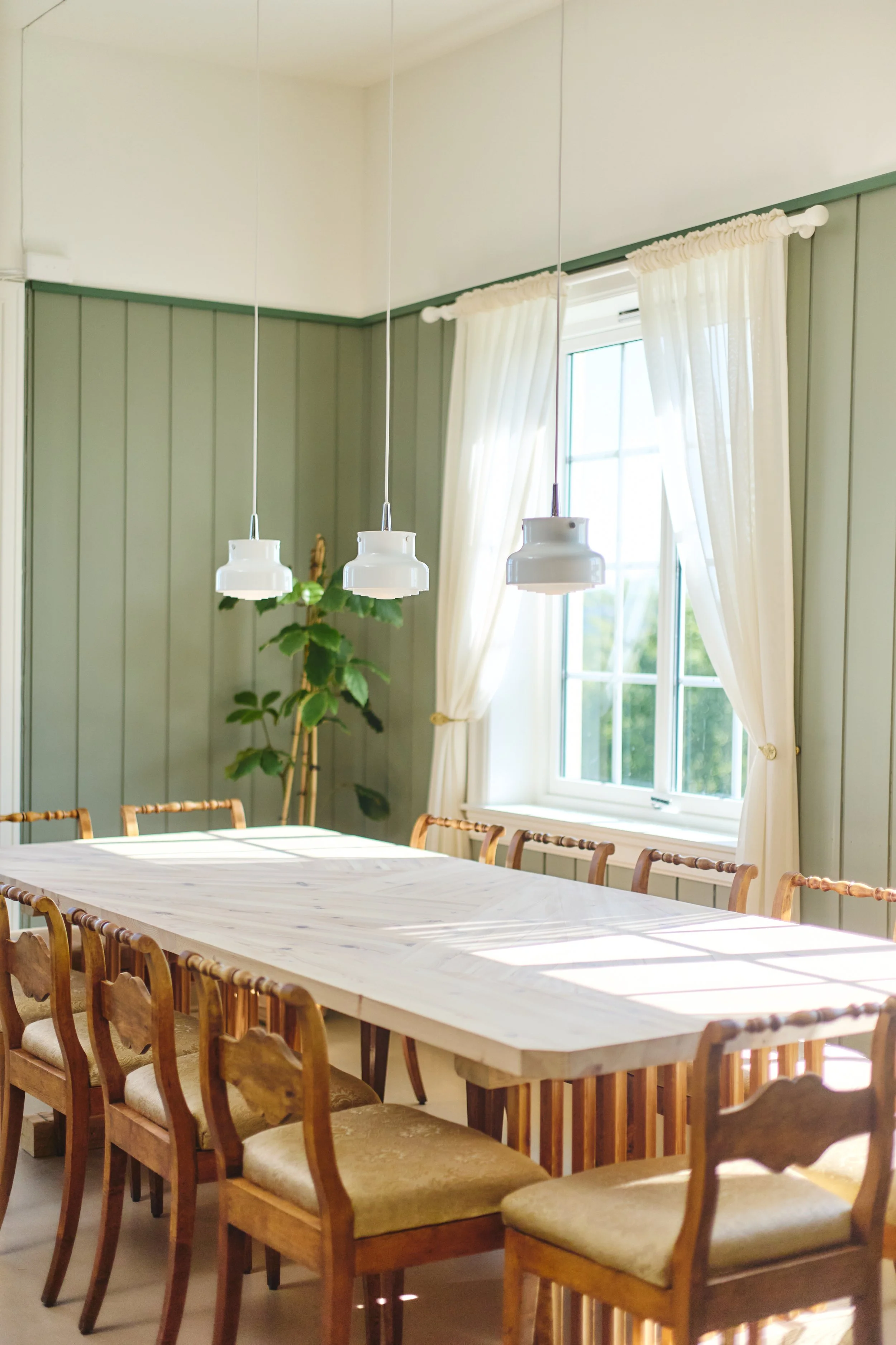 Et lyst rom med et langt spisebord av tre omgitt av elleve stoler i tre med lyse puter. En stor vindu med hvite gardiner slipper inn mye dagslys. Det er en plante i bakgrunnen.
