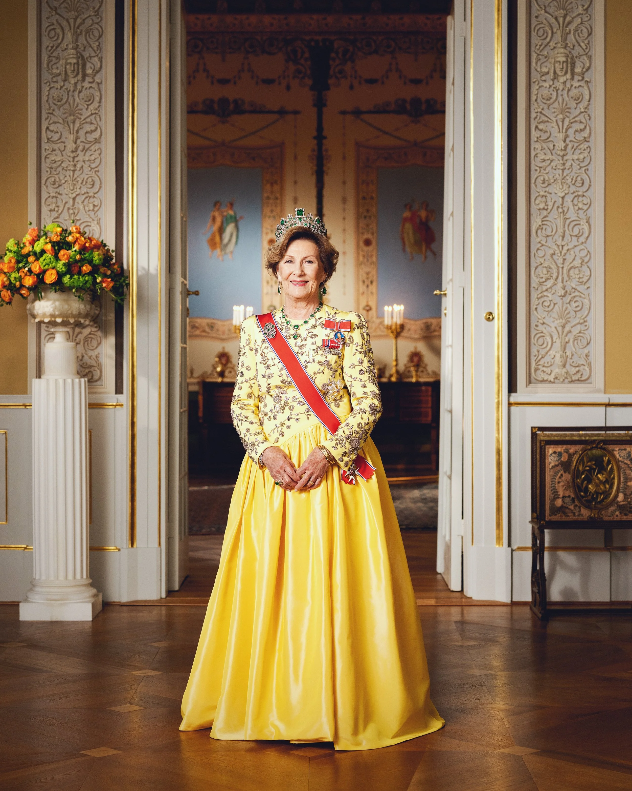 Dronning Sonja oppstilt i elegant salong med gul kjole, diadem og regalia. Bak henne, en oval korridor med malte veggdekorasjoner og fyrlys.