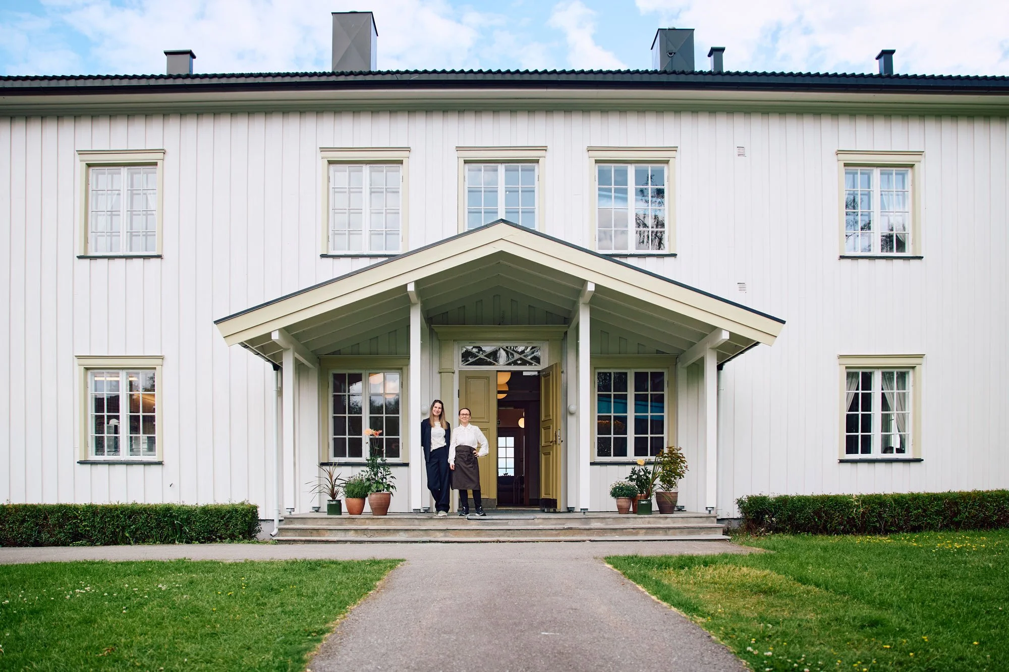To personer står foran inngangen til en hvit trebygning med tre store vinduer og en takterrasse. Det er en grønne plen og en sti som fører opp til trappen.
