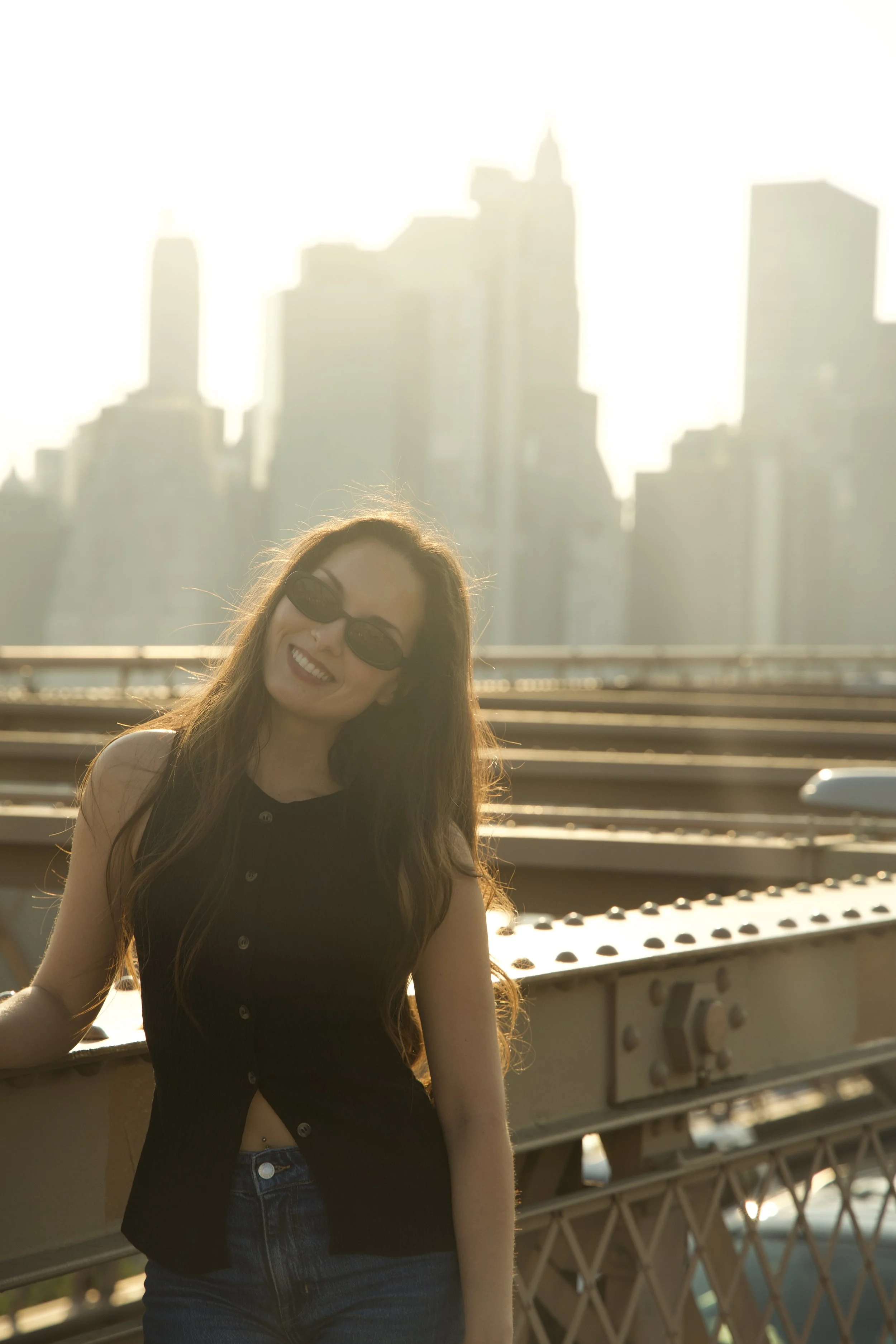 A smiling woman wearing sunglasses and a black sleeveless shirt, standing on a bridge with a city skyline in the background during sunset.