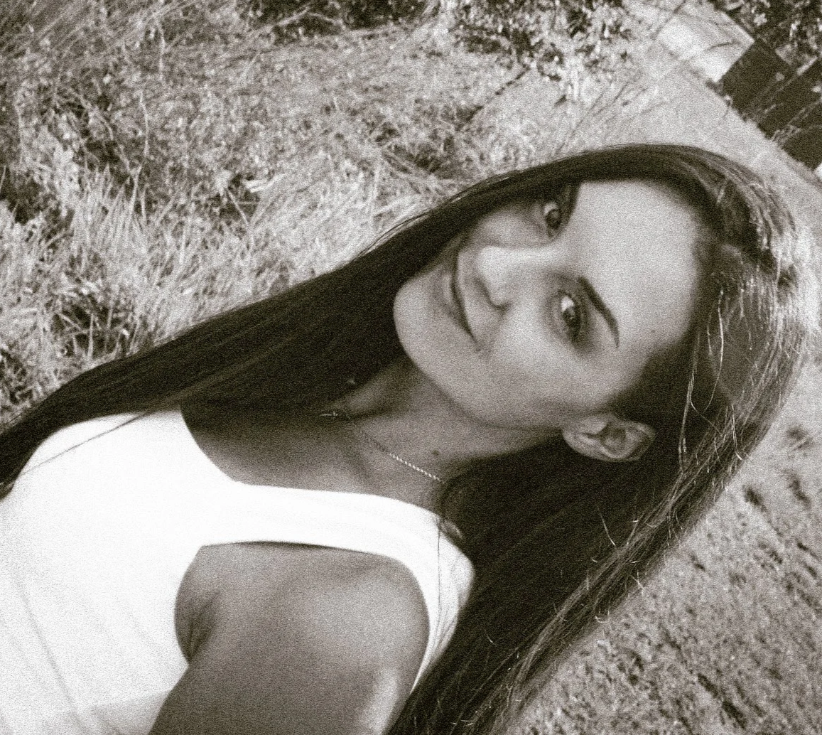 A smiling woman wearing sunglasses and a black sleeveless shirt, standing on a bridge with a city skyline in the background during sunset.