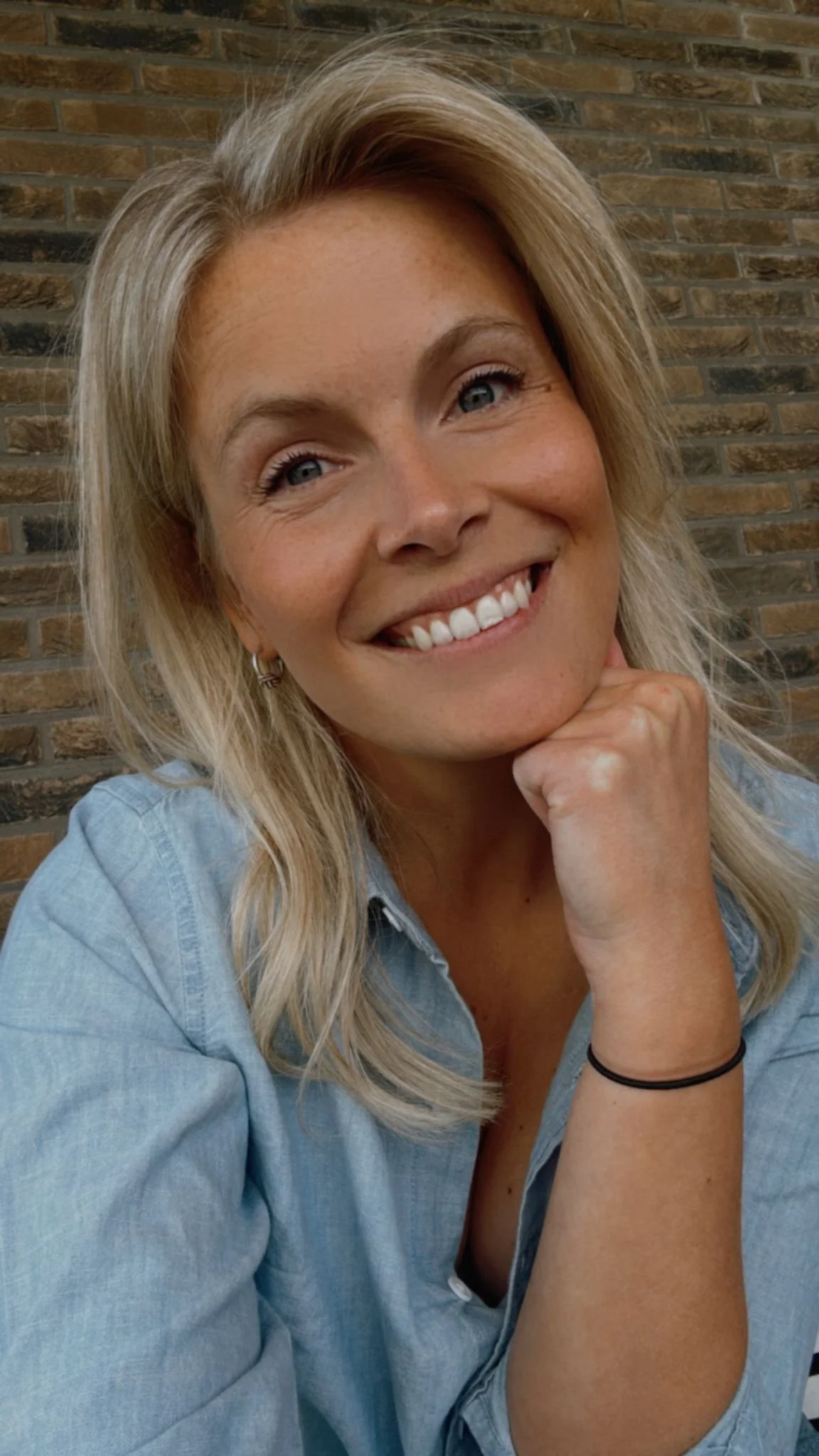 Portret van een glimlachende blonde vrouw met blauwe ogen, zittend voor een muur van bakstenen, rustend op haar hand.