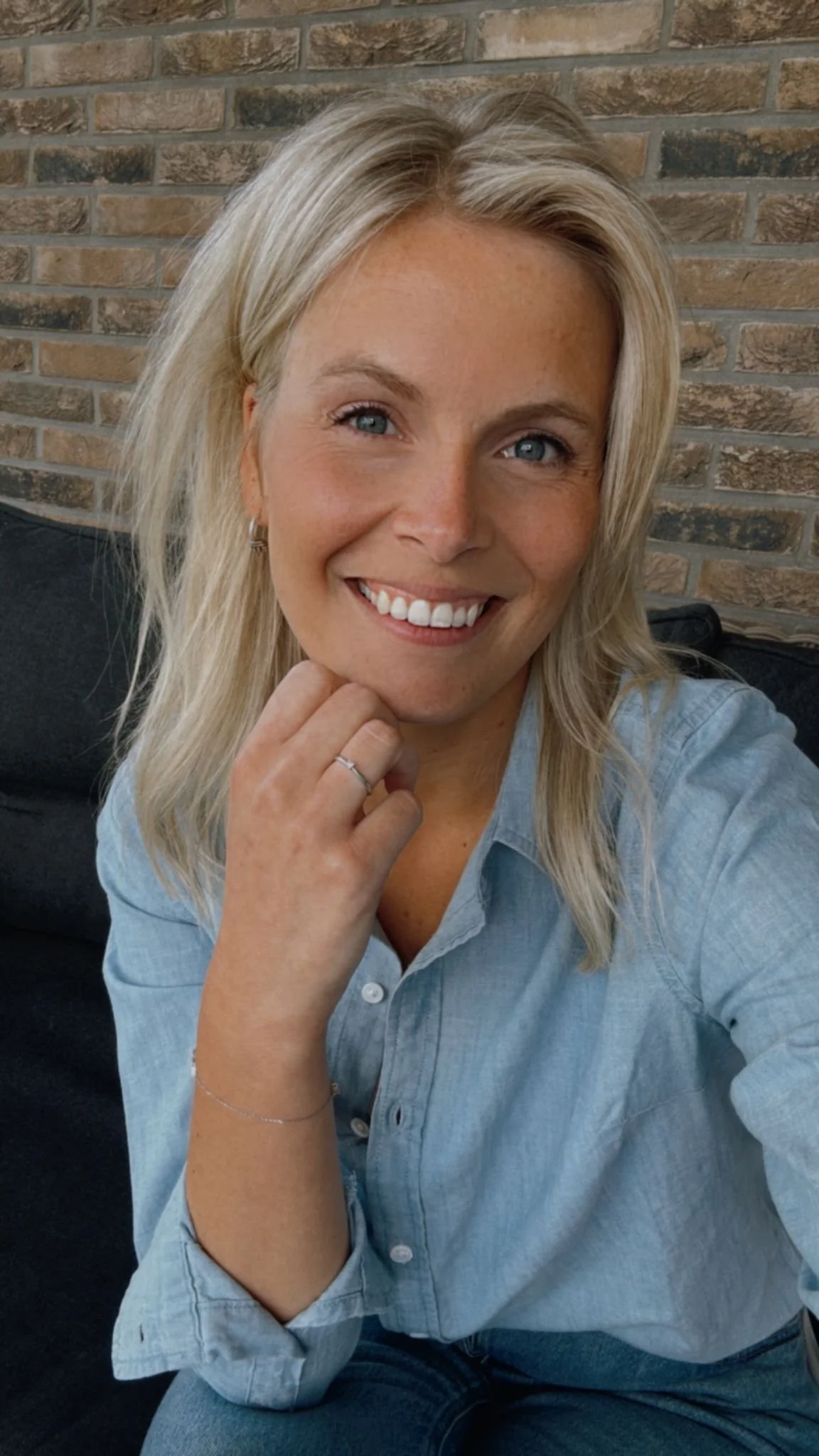 Een glimlachende vrouw met blond haar en blauwe ogen, zittend op een bank voor een bakstenen muur, gekleed in een lichtblauw overhemd.
