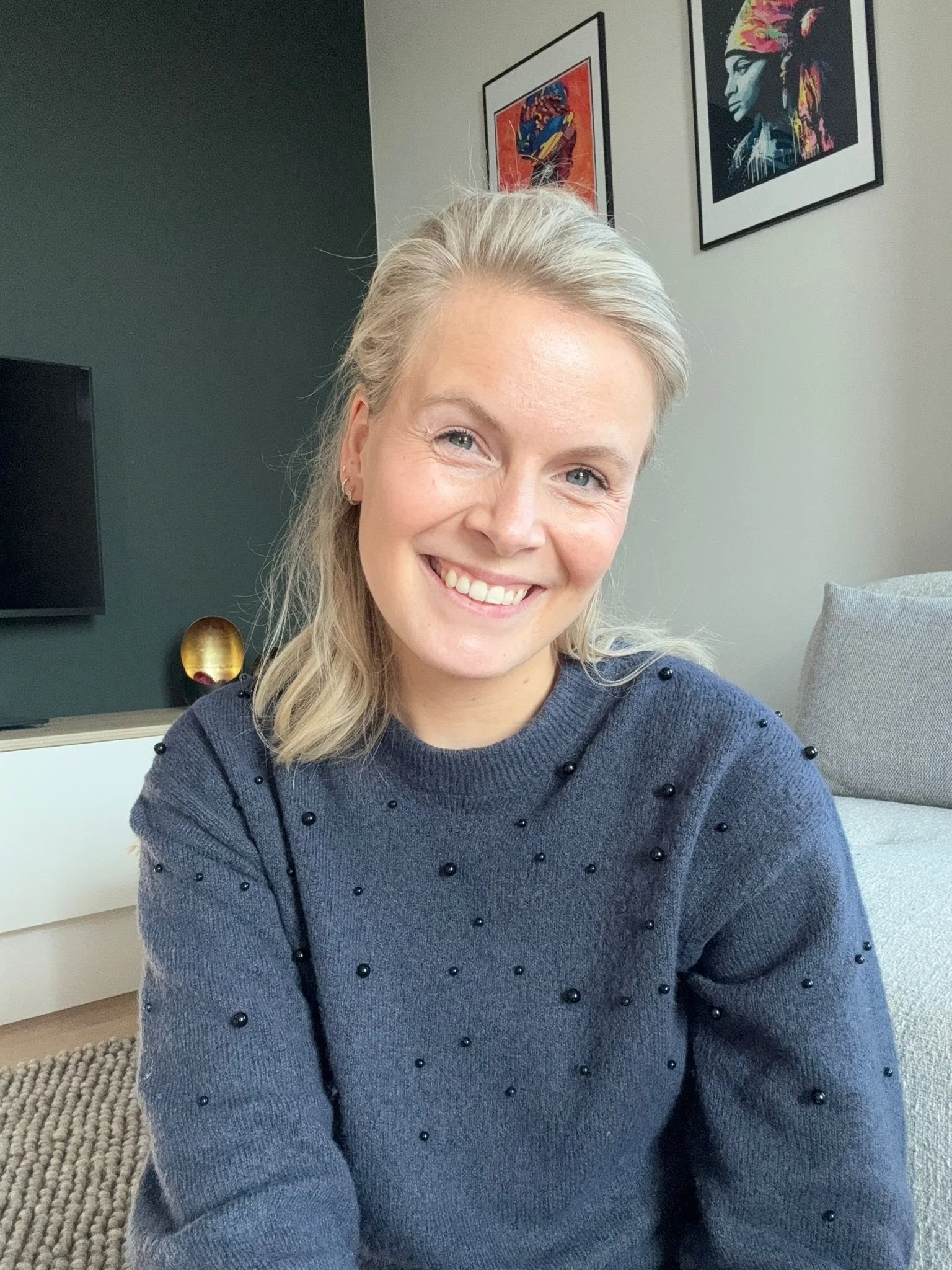Een lachende vrouw met blond haar, zittend in een woonkamer, gekleed in een donkergrijze trui met zwarte parels, op de achtergrond kunstwerken aan de muur en een televisie.