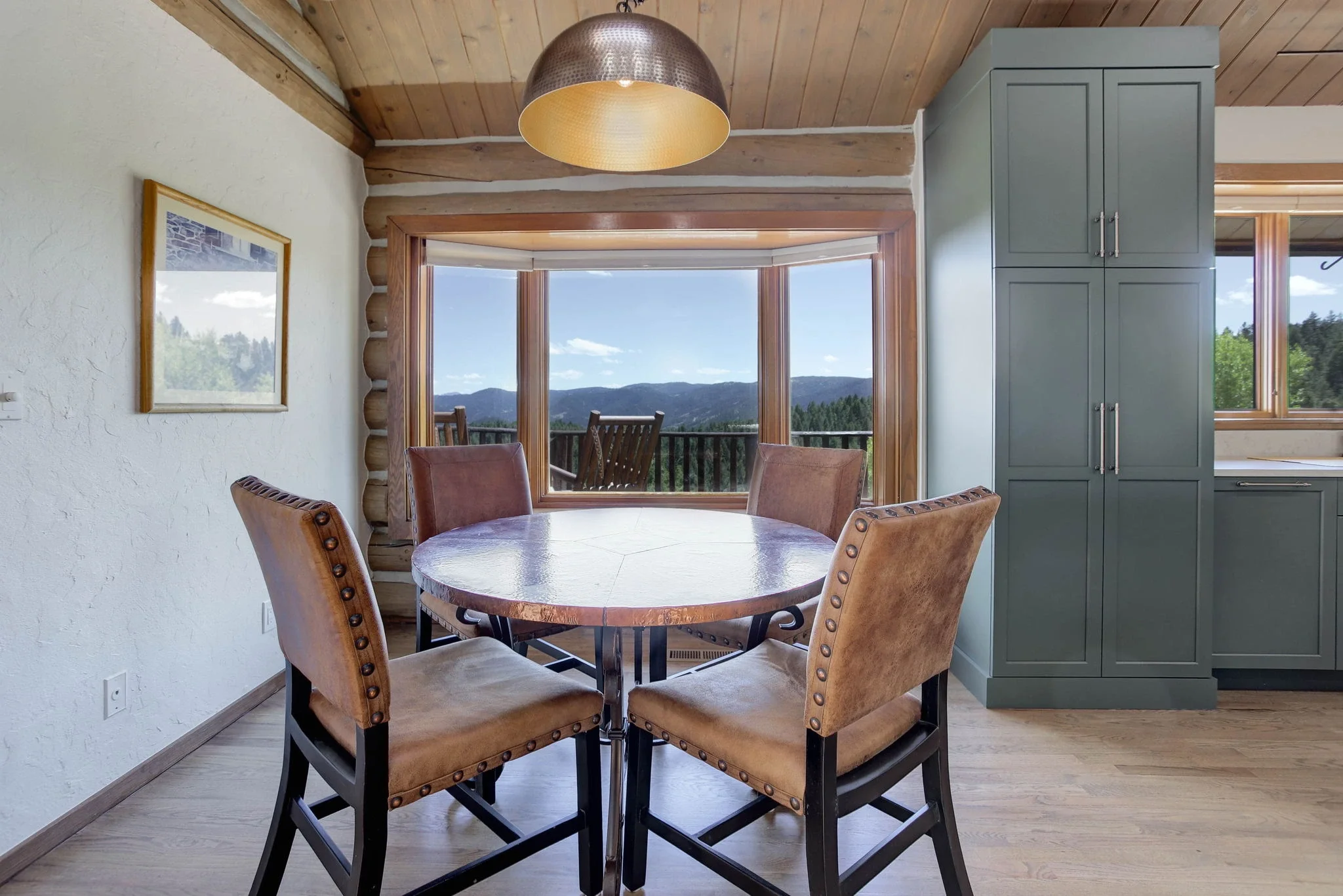 A dining room with a round table, four leather chairs, a large window with a mountain view, a green cabinet, and a ceiling light.