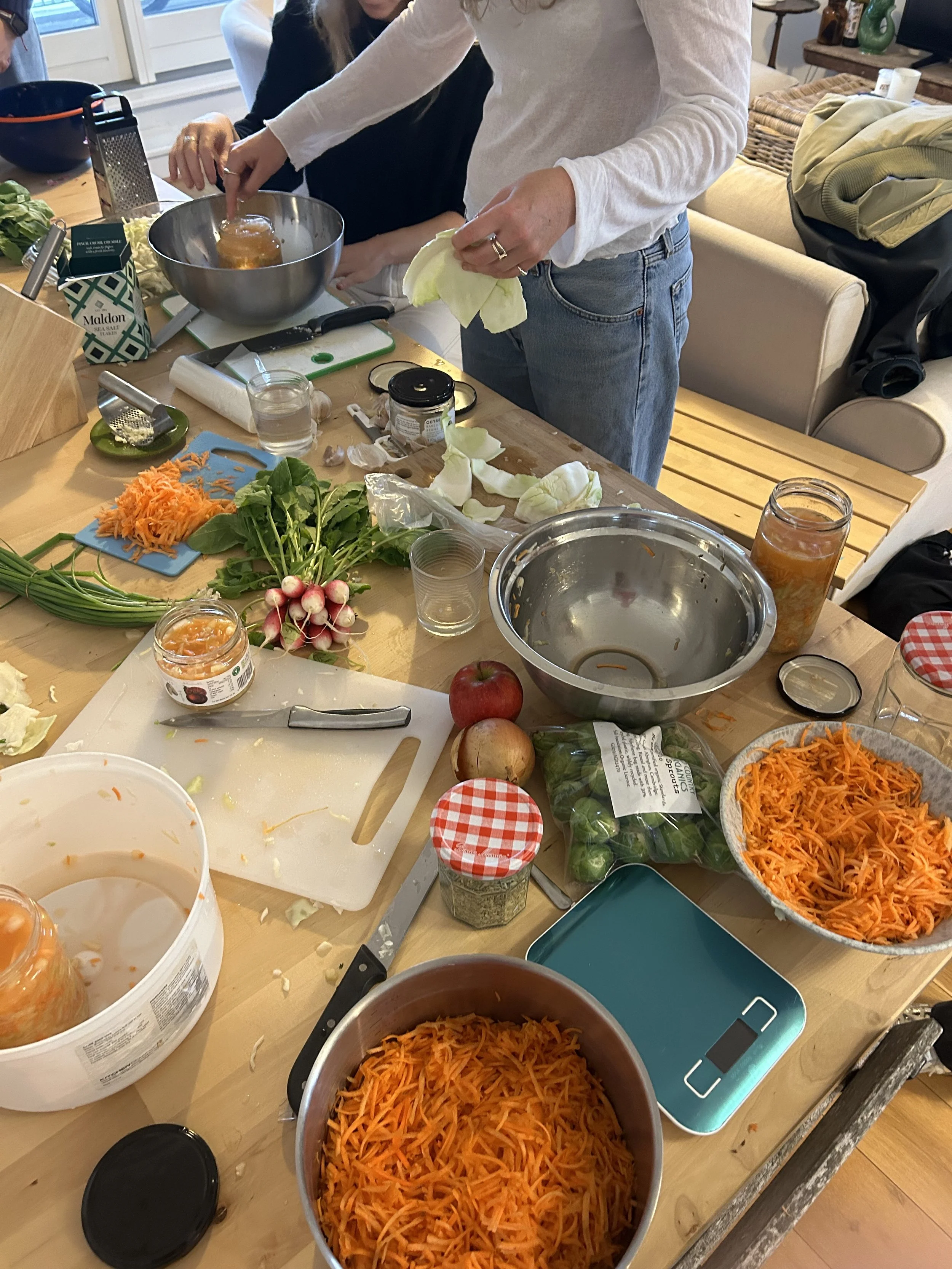People preparing food in a kitchen, with shredded carrots, radishes, onions, and various jars of ingredients on the table, including jars of preserved vegetables and seasonings.