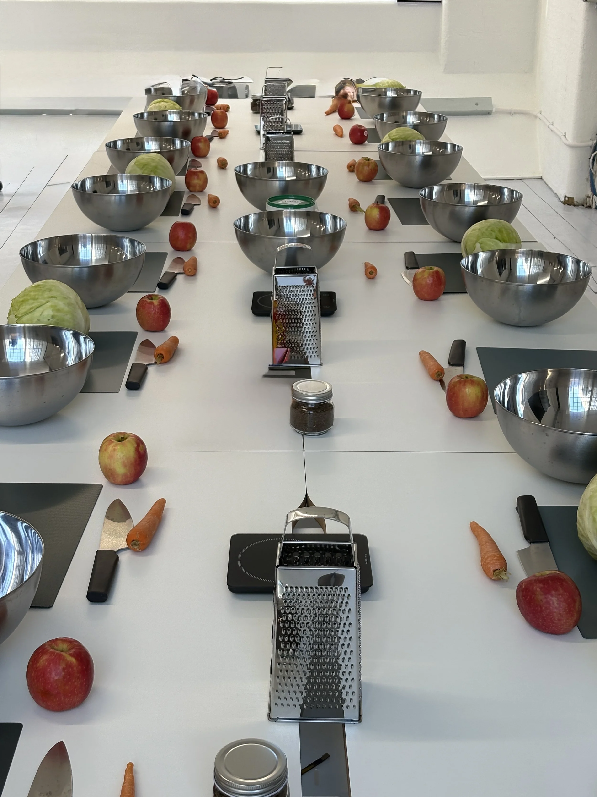 A long white kitchen worktable with apples, carrots, and hazelnuts arranged next to stainless steel bowls and graters, with kitchen gadgets in the background.
