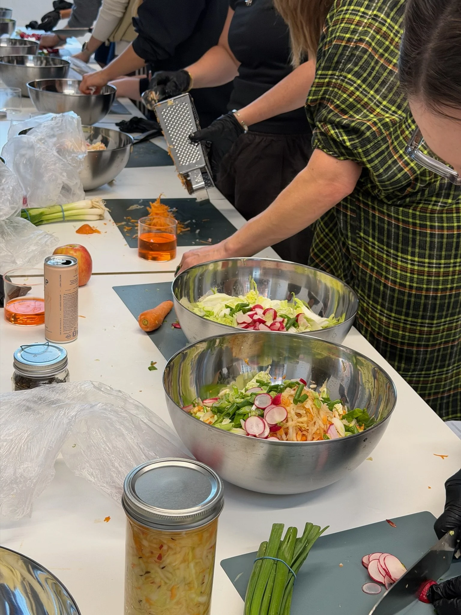 People preparing salads at a cooking class, with bowls of chopped vegetables, carrots, radishes, green onions, and glasses of drinks on the table.