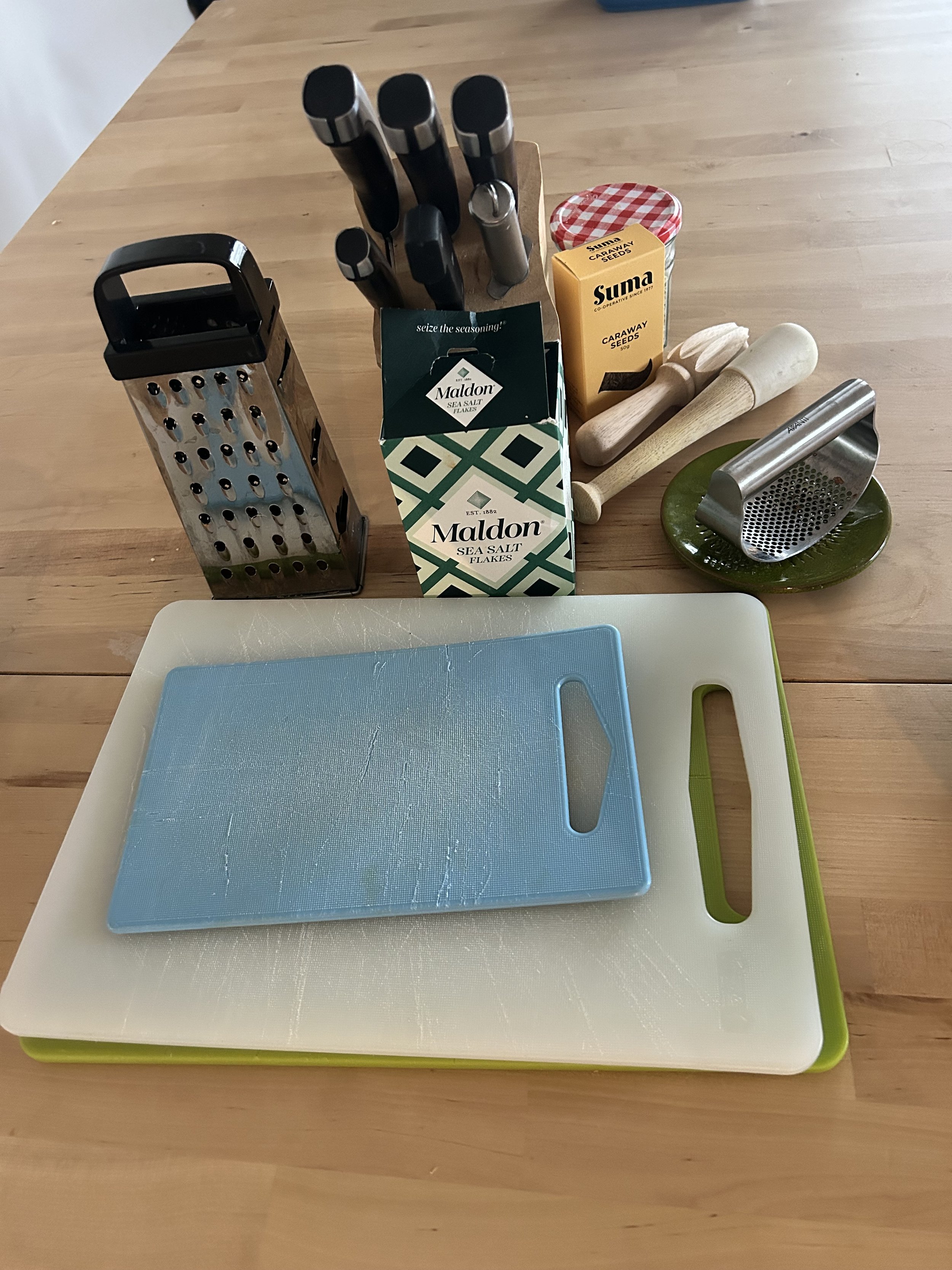 A kitchen countertop with a blue cutting board on a white and green tray, a cheese grater, a knife block with knives, two wooden mallets, a jar of Sumac seeds, and a box of Maldon sea salt flakes.