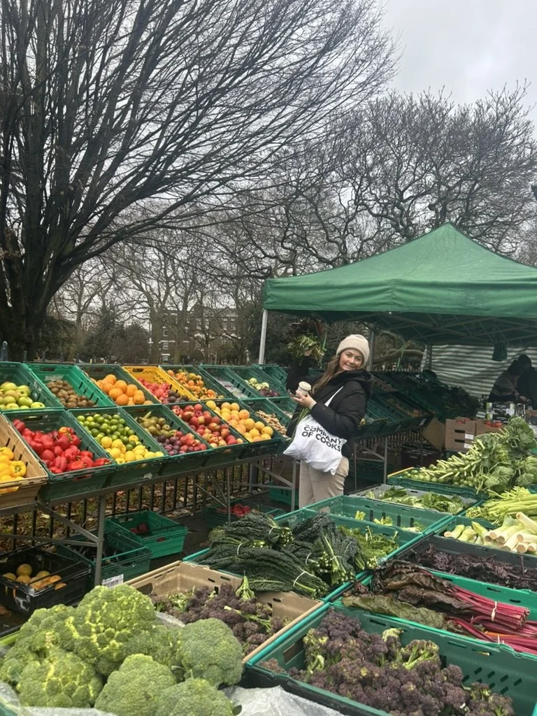 A woman at a farmers market stall full of fresh vegetables and fruits, holding a cup and shopping bag, under a green canopy on a cloudy day with leafless trees in the background.