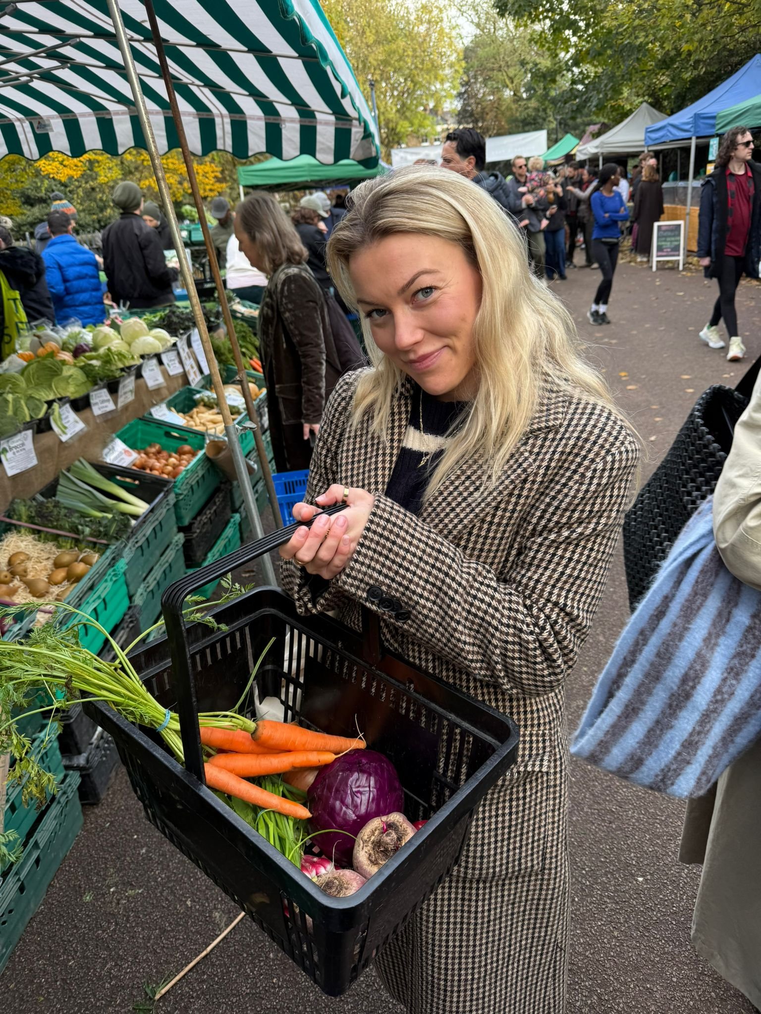 A woman with blonde hair shopping at an outdoor market, holding a black shopping basket filled with carrots, radicchio, onions, mushrooms, and other vegetables, with market stalls and other shoppers in the background.