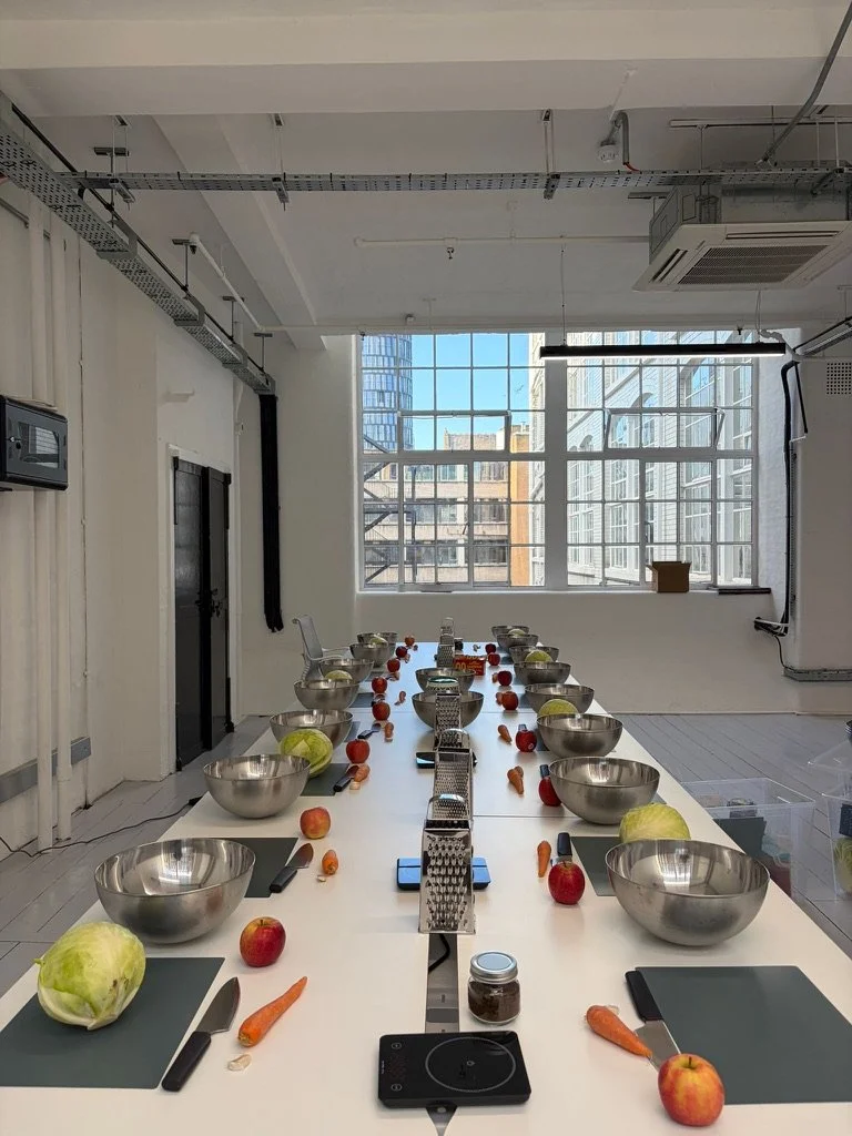 A modern, industrial-style kitchen with a long white dining table set for a meal or cooking class. The table has bowls, apples, carrots, and small graters arranged on it, with black placemats. Large windows in the background let in natural light and reveal city buildings outside.
