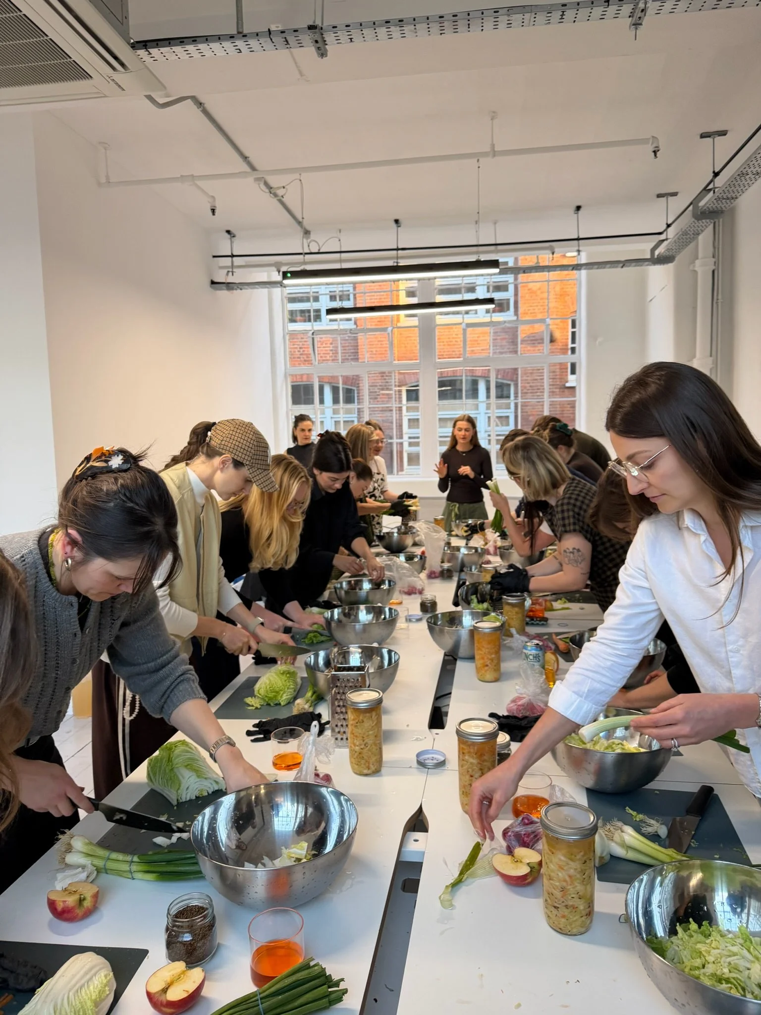 Group of people participating in a culinary or food preparation class in a bright room with large windows, preparing ingredients on a long table.