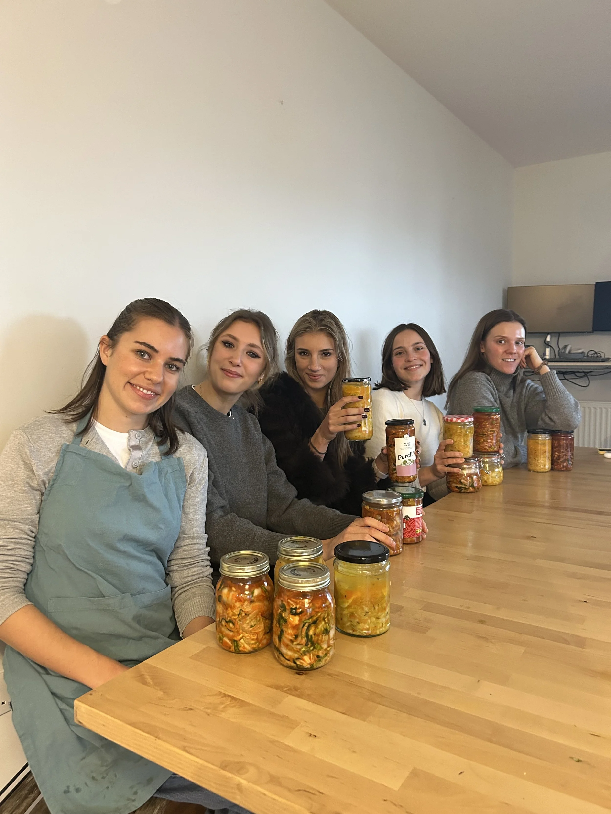 Five women sitting at a wooden table with jars of homemade preserved vegetables, smiling at the camera.
