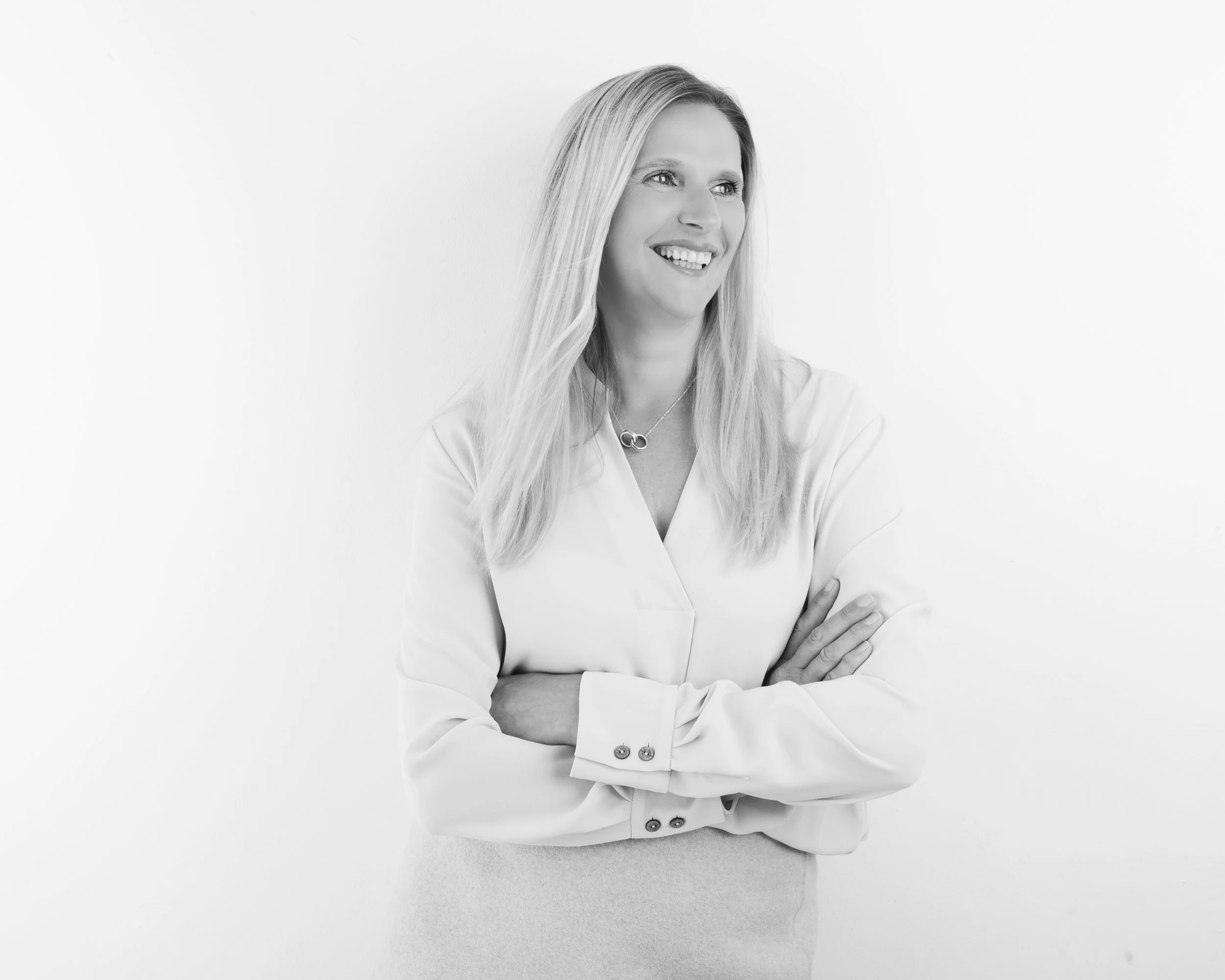 A woman with long blonde hair smiling slightly, standing with arms crossed against a plain white background, wearing a blazer and a necklace.