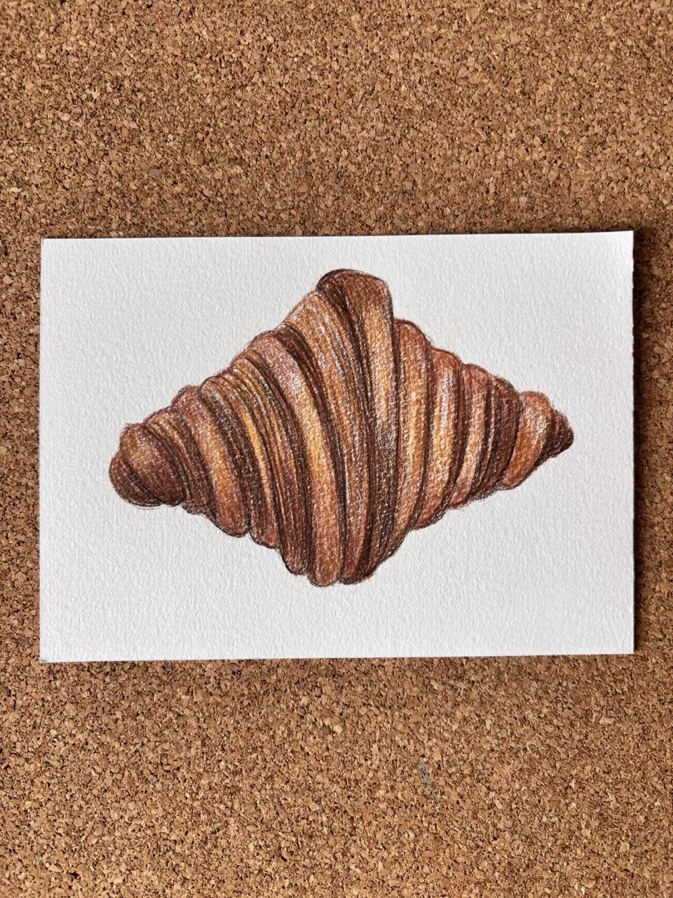 Colorful drawing of a croissant on a white textured paper, placed on a cork surface.