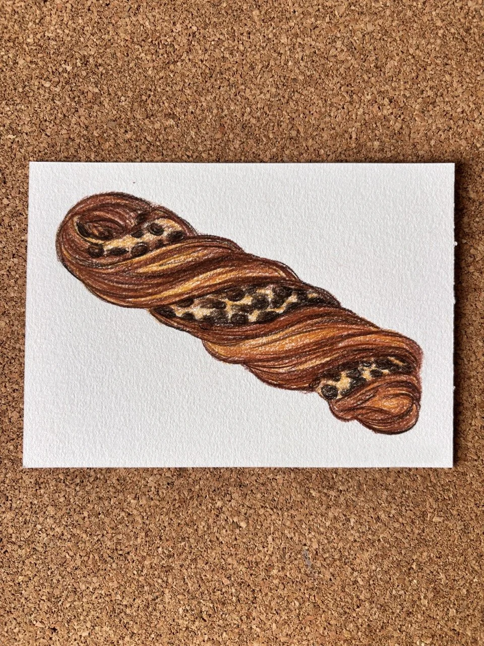A detailed illustration of a chocolate croissant with visible layers and chocolate filling, placed on a white piece of paper on a corkboard background.