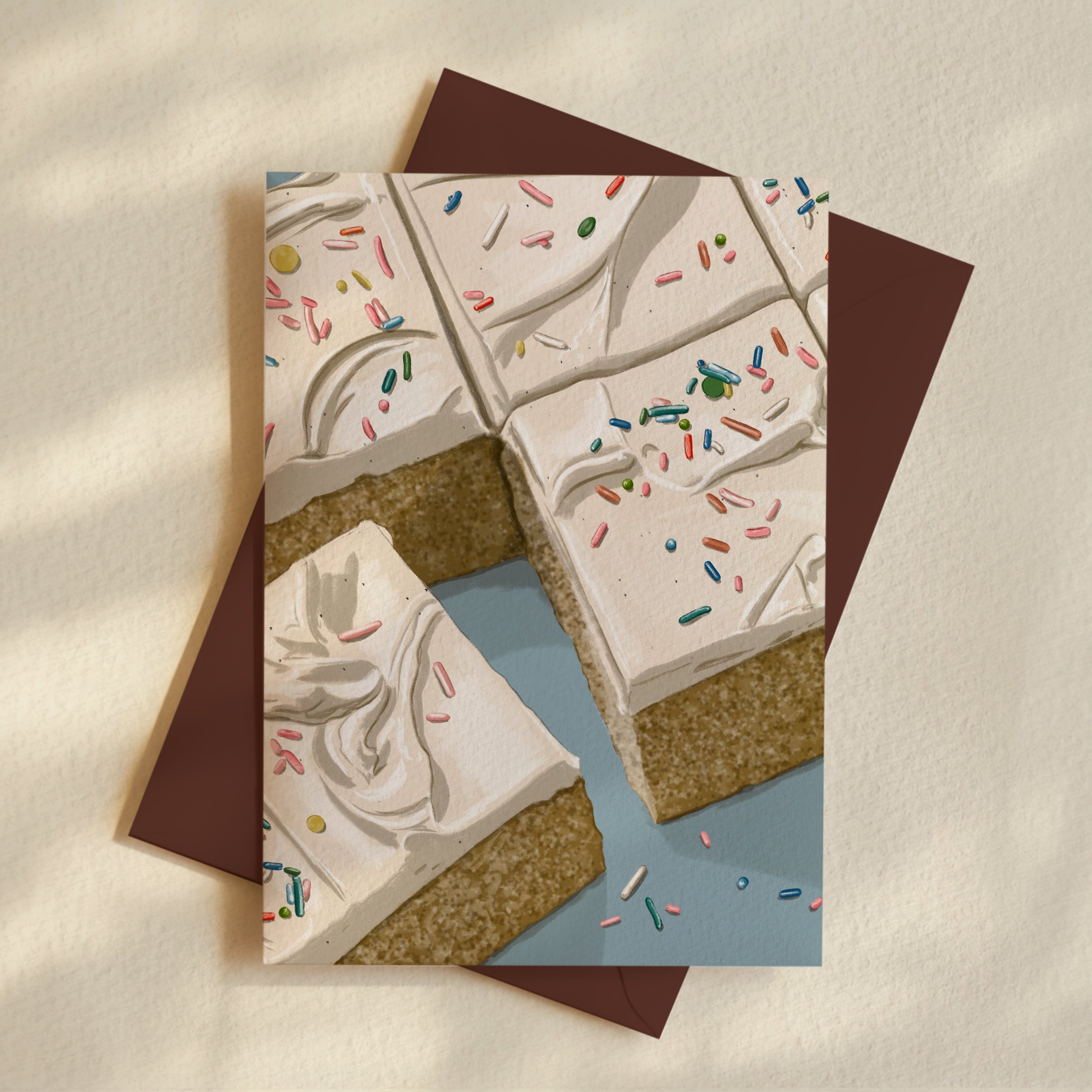 A birthday card with a white frosting design decorated with colorful sprinkles, resting on a beige surface with a brown envelope behind it.