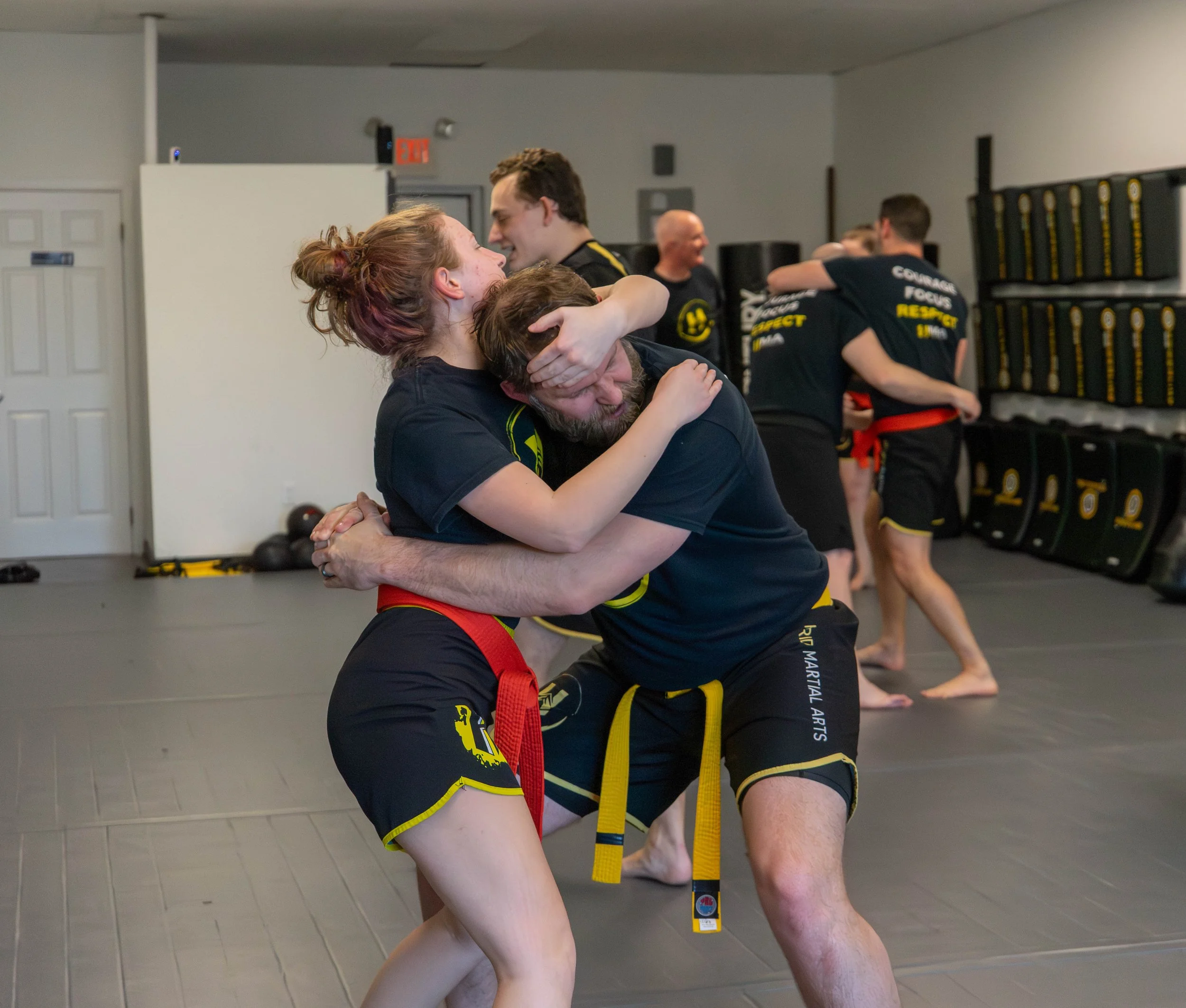 Two martial artists practicing a grappling move in a martial arts gym, with others observing in the background.