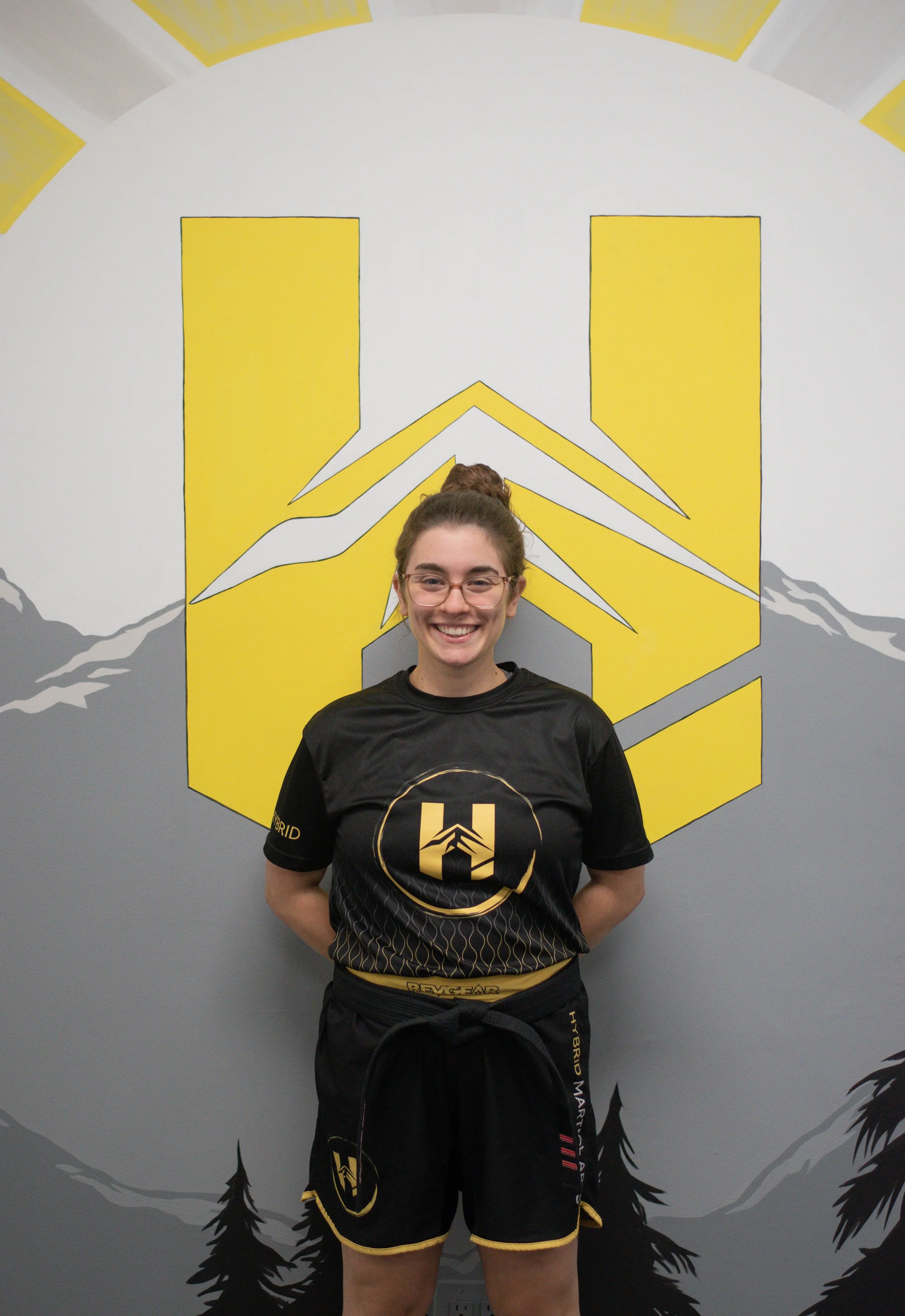 A smiling woman with glasses, wearing a black martial arts uniform with yellow accents, standing in front of a wall with yellow and gray design elements and a graphic of mountains and trees.
