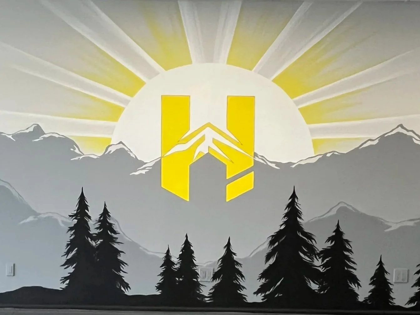 Wall mural featuring a sunrise or sunset behind mountain ranges, with black pine trees in the foreground and a yellow logo with mountains and bold shapes in front.