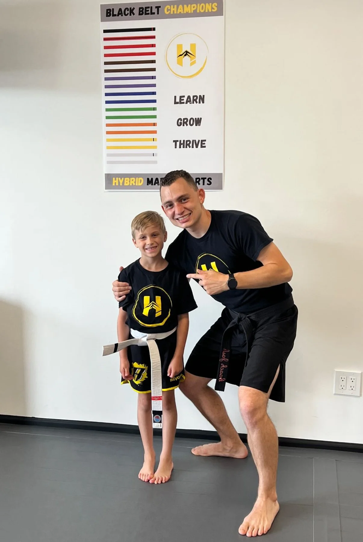 A young boy and an adult male, both wearing black martial arts uniforms with yellow and black logos, pose with smiles in a martial arts studio. The boy has a white belt, and the man points to him with a big smile. There is a poster on the wall behind them titled "Black Belt Champions" with various colored stripes and the words "Learn, Grow, Thrive."