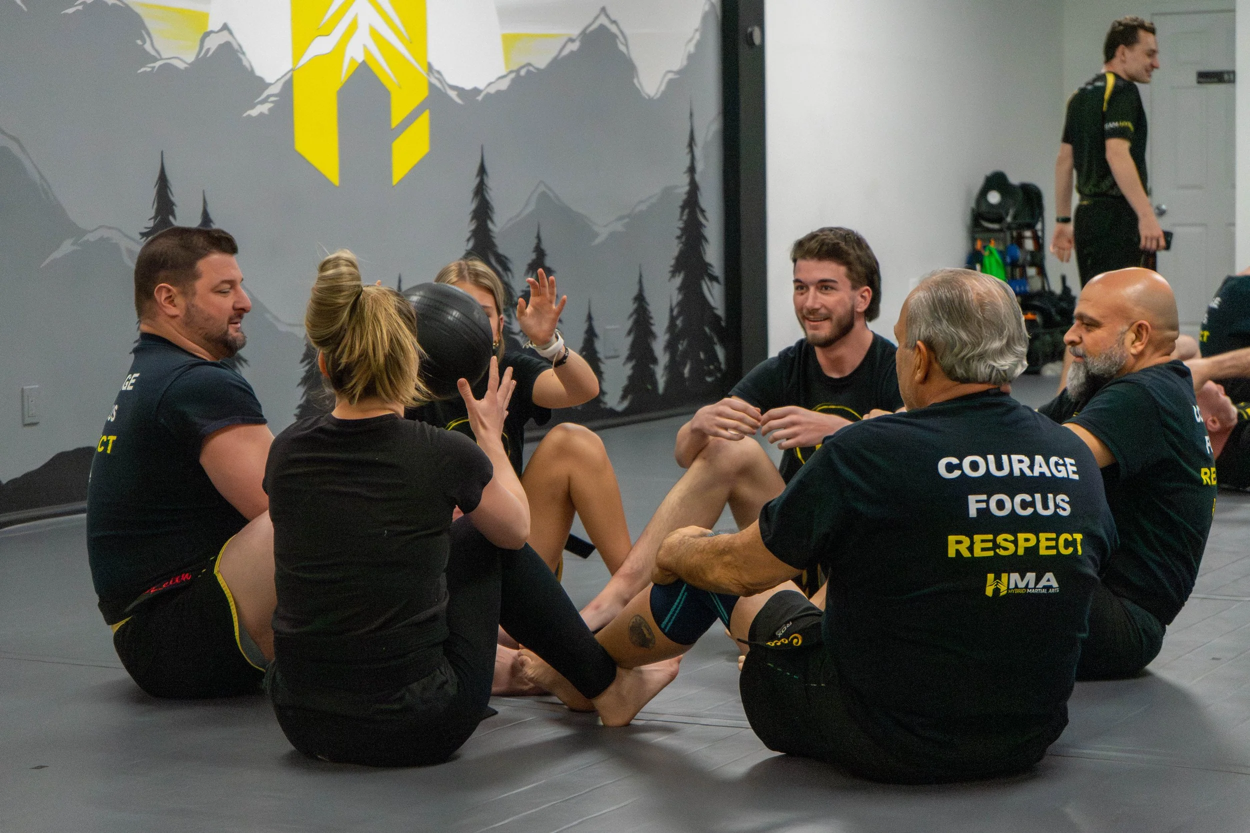 Group of people sitting on the floor in a martial arts gym, engaging in a training session or demonstration. They are wearing black training attire with some words printed on the back, such as 'COURAGE', 'FOCUS', 'RESPECT'. The gym has a mural of mountains, trees, and a logo on the wall.